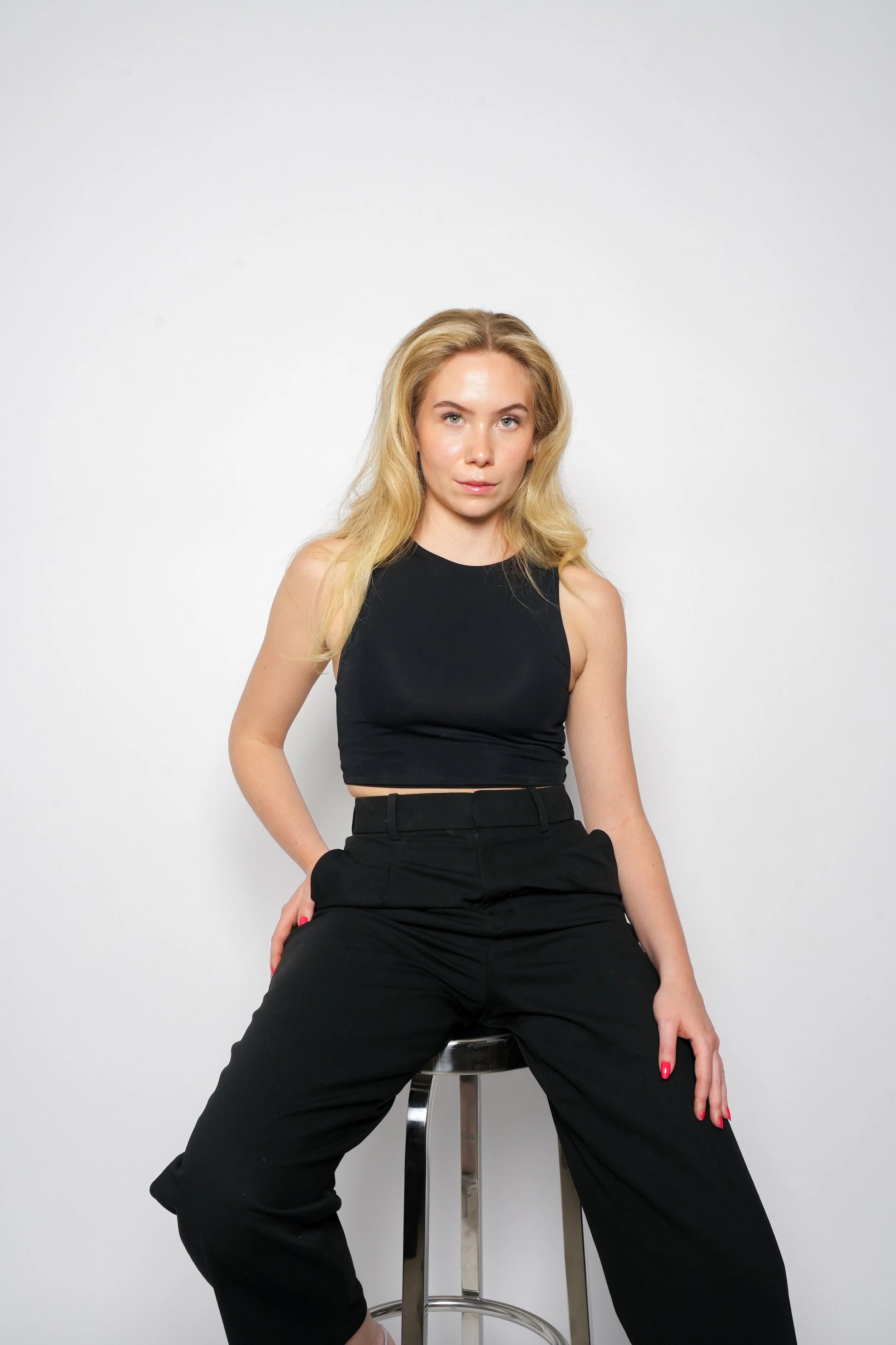 Kvindelig person sidder på en barstol iført sort top og bukser mod en hvid baggrund.