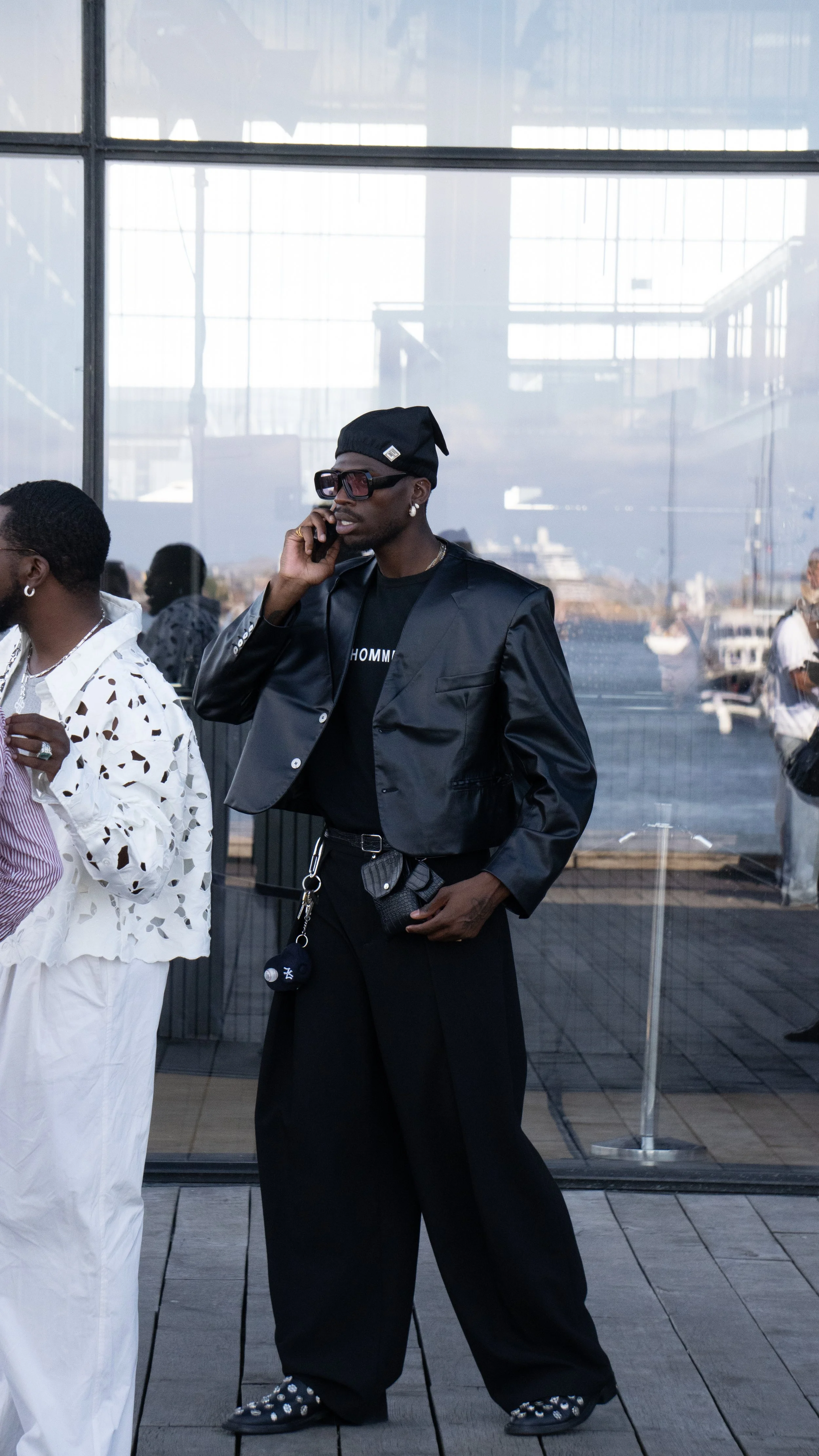 Person i sort jakke og bredel, sort hat, store solbriller, taler i telefon, står foran glasvæg med udsigt over havn med både.