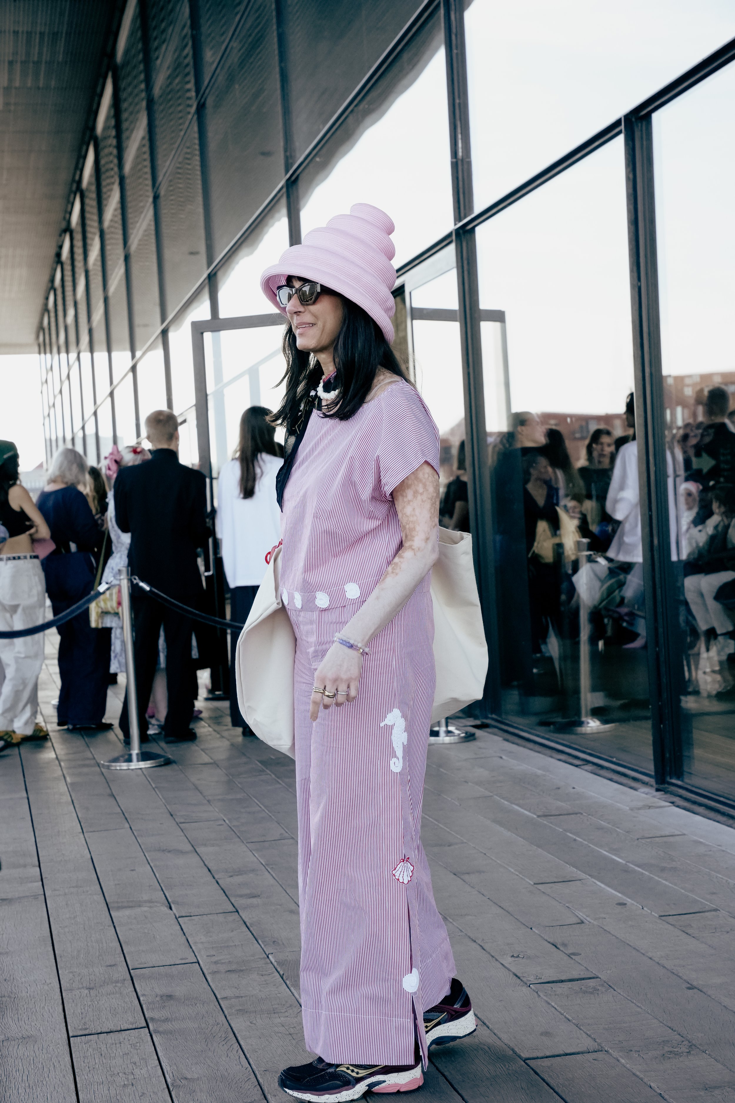 En person iført en lyserød og hvid stribet kjole med motiver af havdyr, en lyserød hat, solbriller og sneakers, der står udenfor ved en glasbygning med mennesker i baggrunden.