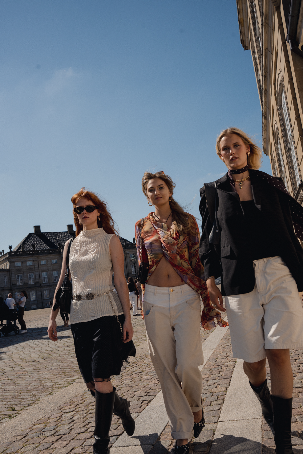 Tre unge kvinder går på gaden i solskinsvejr, iført moderne og stilfuldt tøj, med gamle bygninger i baggrunden.
