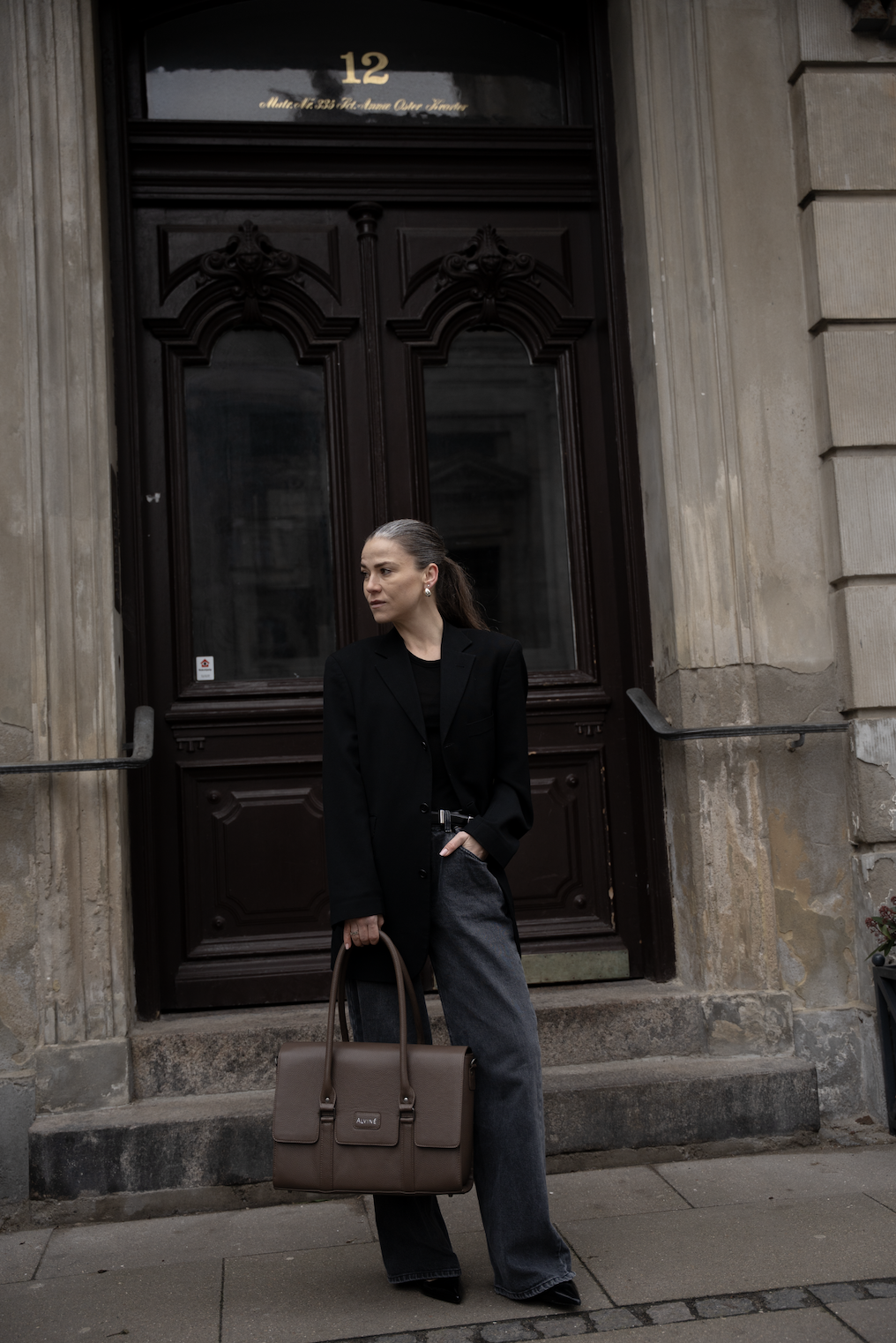 En stilfuld kvinde står foran en stor, antik dør i en bygning i København. Hun bærer en sort blazer, løse grå bukser, har en stor brunt håndtaske og hendes hår er trukket tilbage.