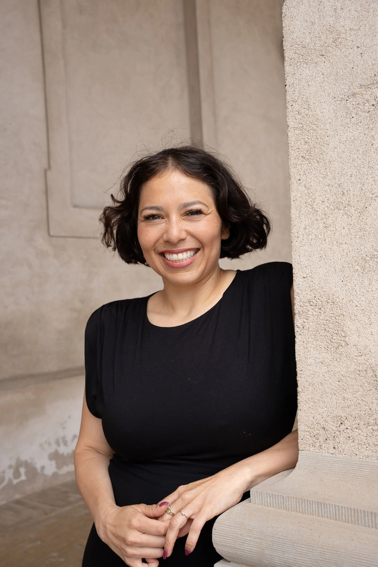 En glad kvinde med kort sort hår smiler og står udendørs ved en væg i en sort kjole.