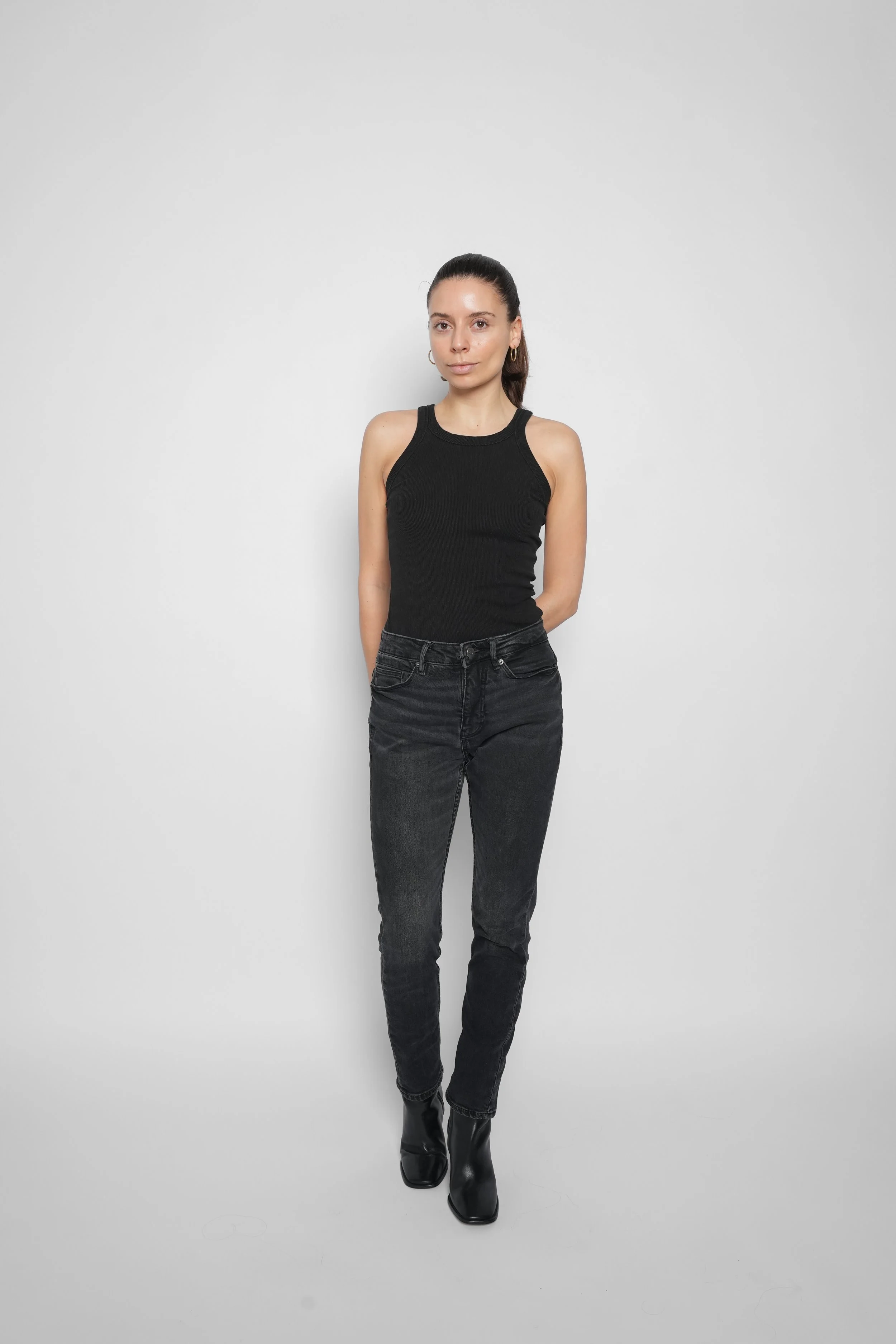 En kvinde står mod en hvid væg, iført sort tanktop, sorte bukser og sorte støvler.