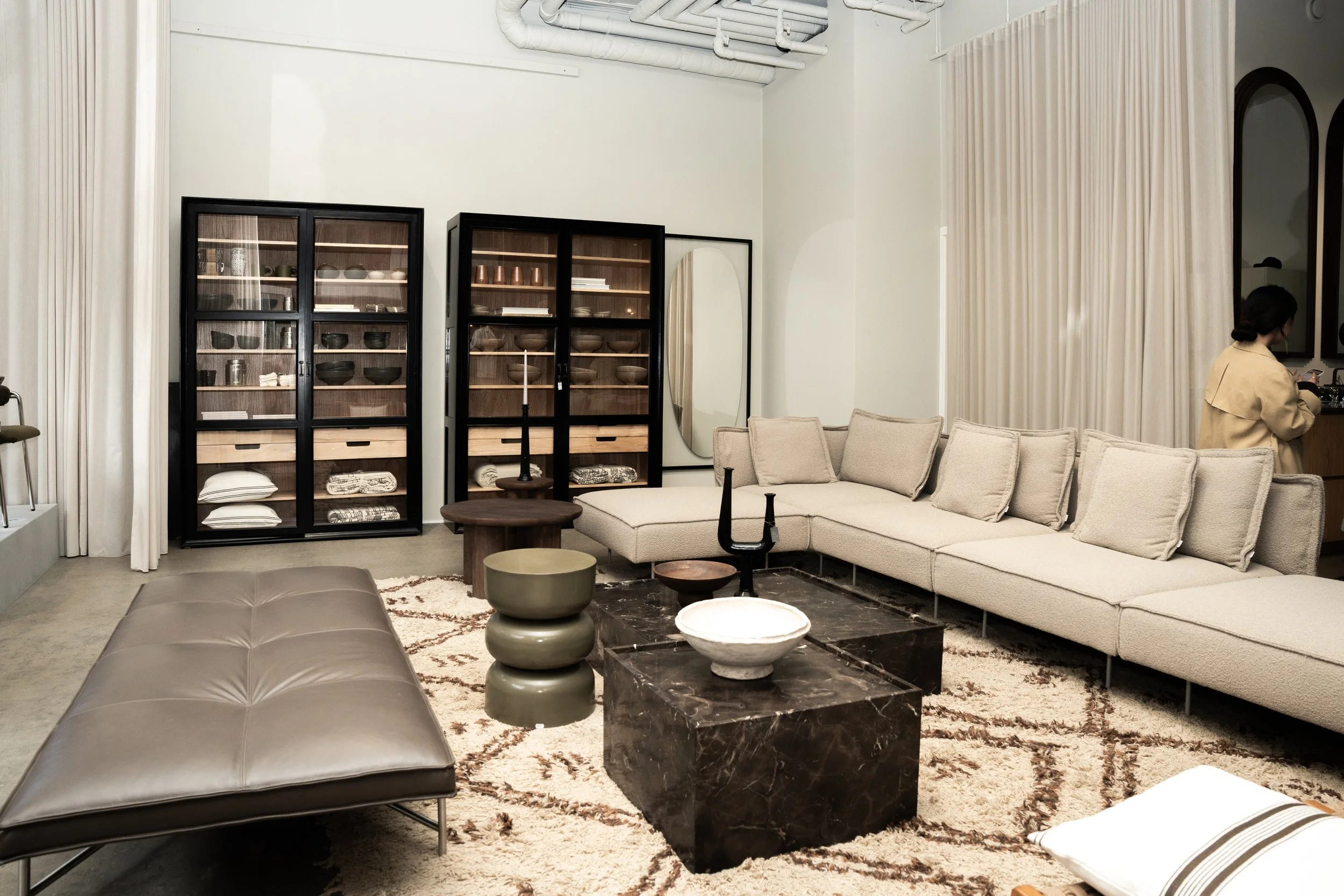 Moderne stue med beige sofa, sort hylder med keramik og tekstiler, spisebord med kandelabre, tæppe med mønster, og en person i beige jakke ved barområdet.
