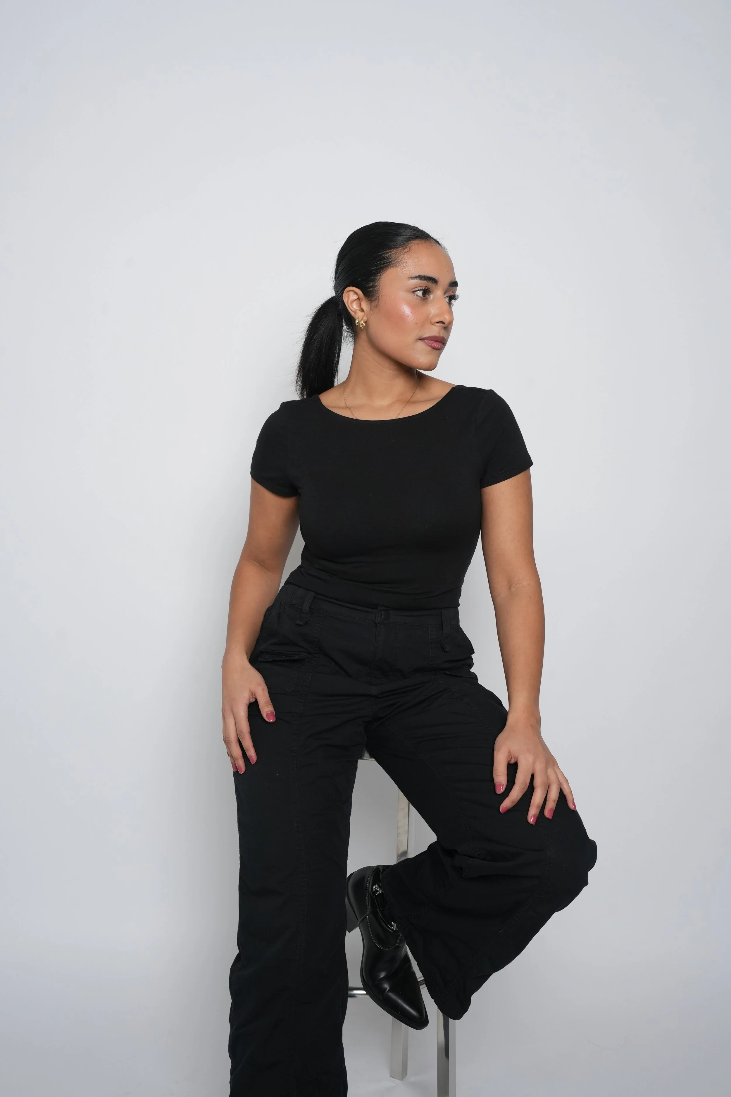 En kvinde med sort hår i hestehale, iført sort t-shirt og sorte bukser, sidder på en stol mod en hvid væg.