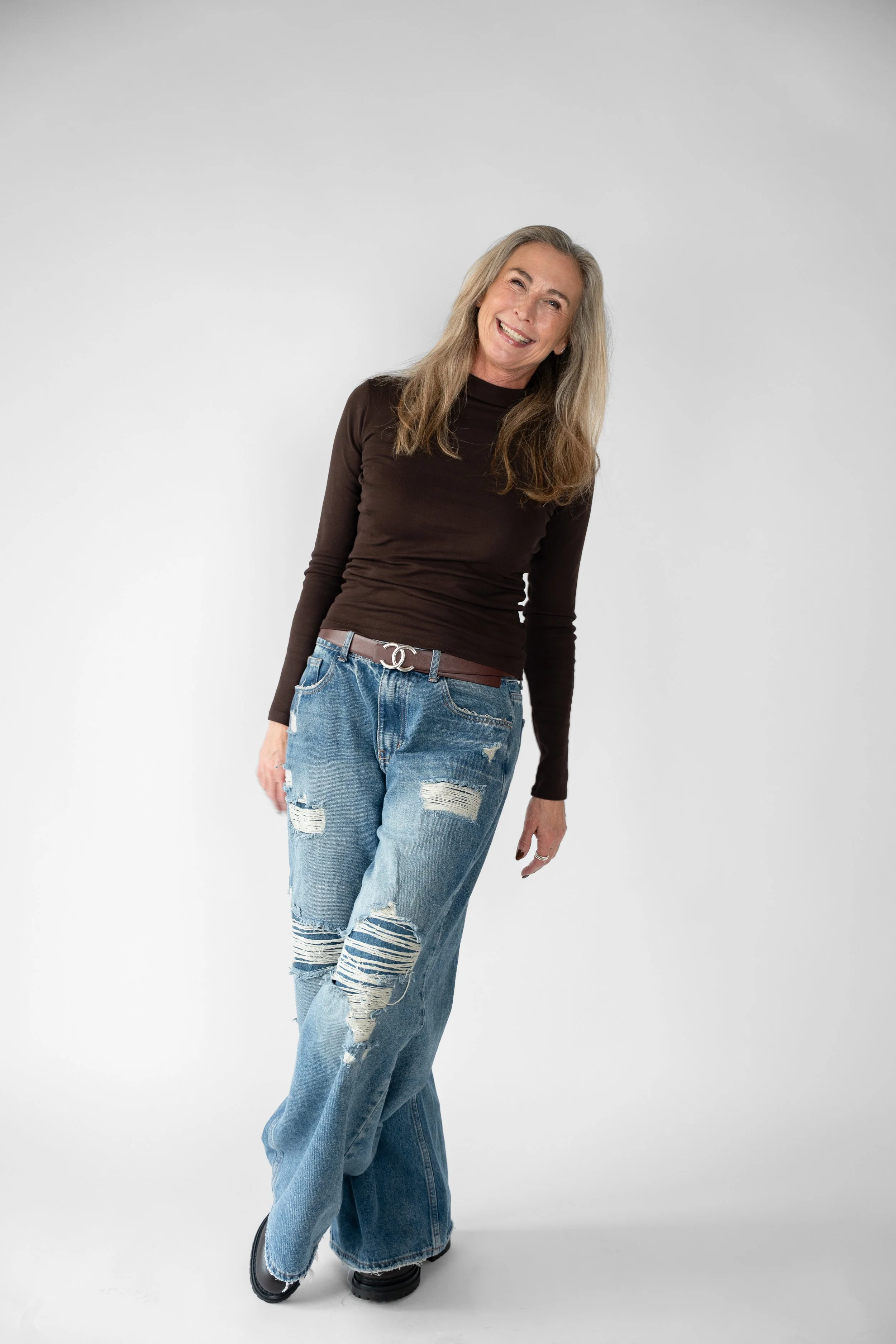Kvinde med langt, blondt hår, iført en sort langærmet top og slidte, store bukser, står mod en hvid baggrund og smiler.