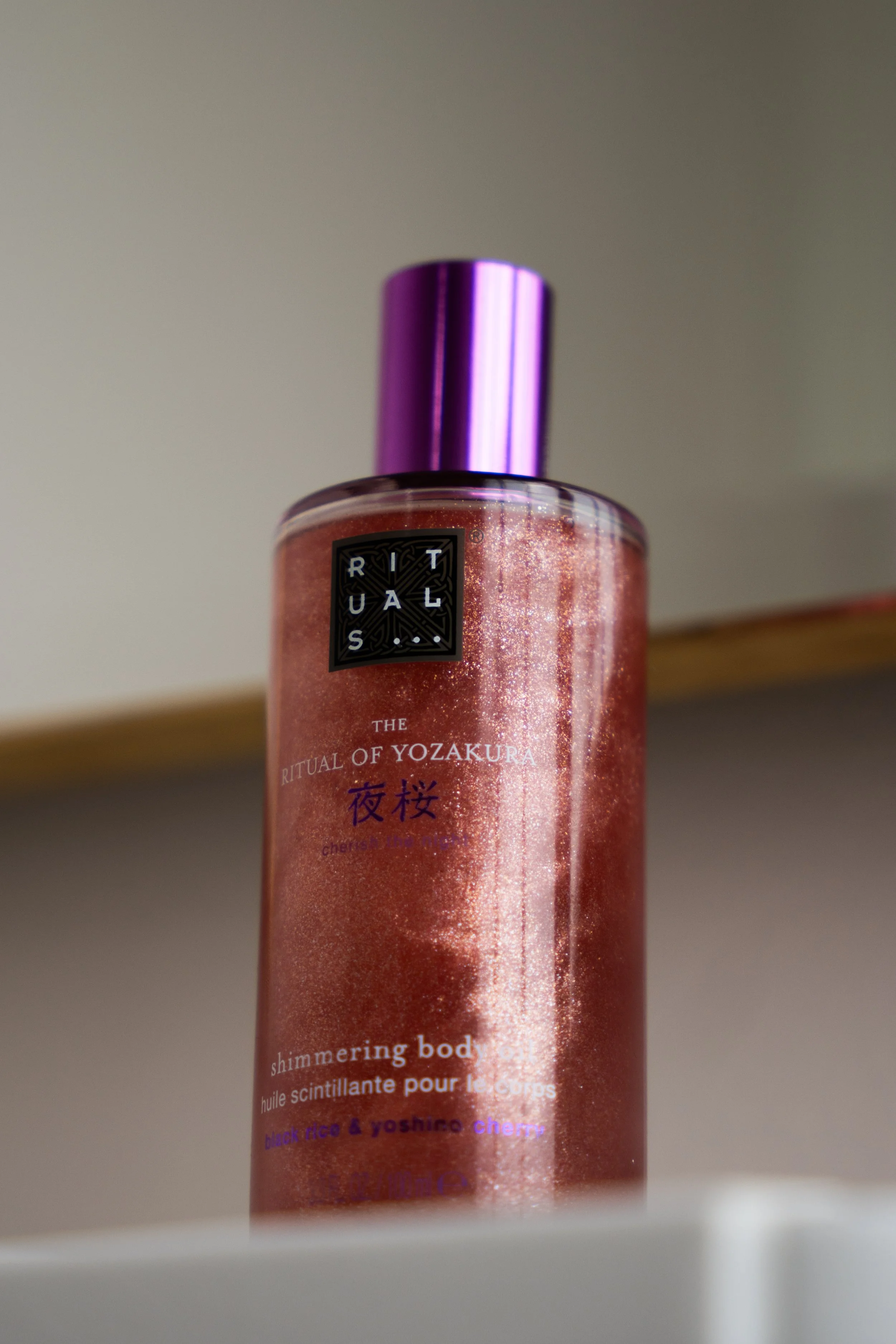 En røgfarvet body mist flaske med lilla låg, mærket 'Rituals The Ritual of Yozakura' med tekst om skinnende krop, og dækker af glimmer.