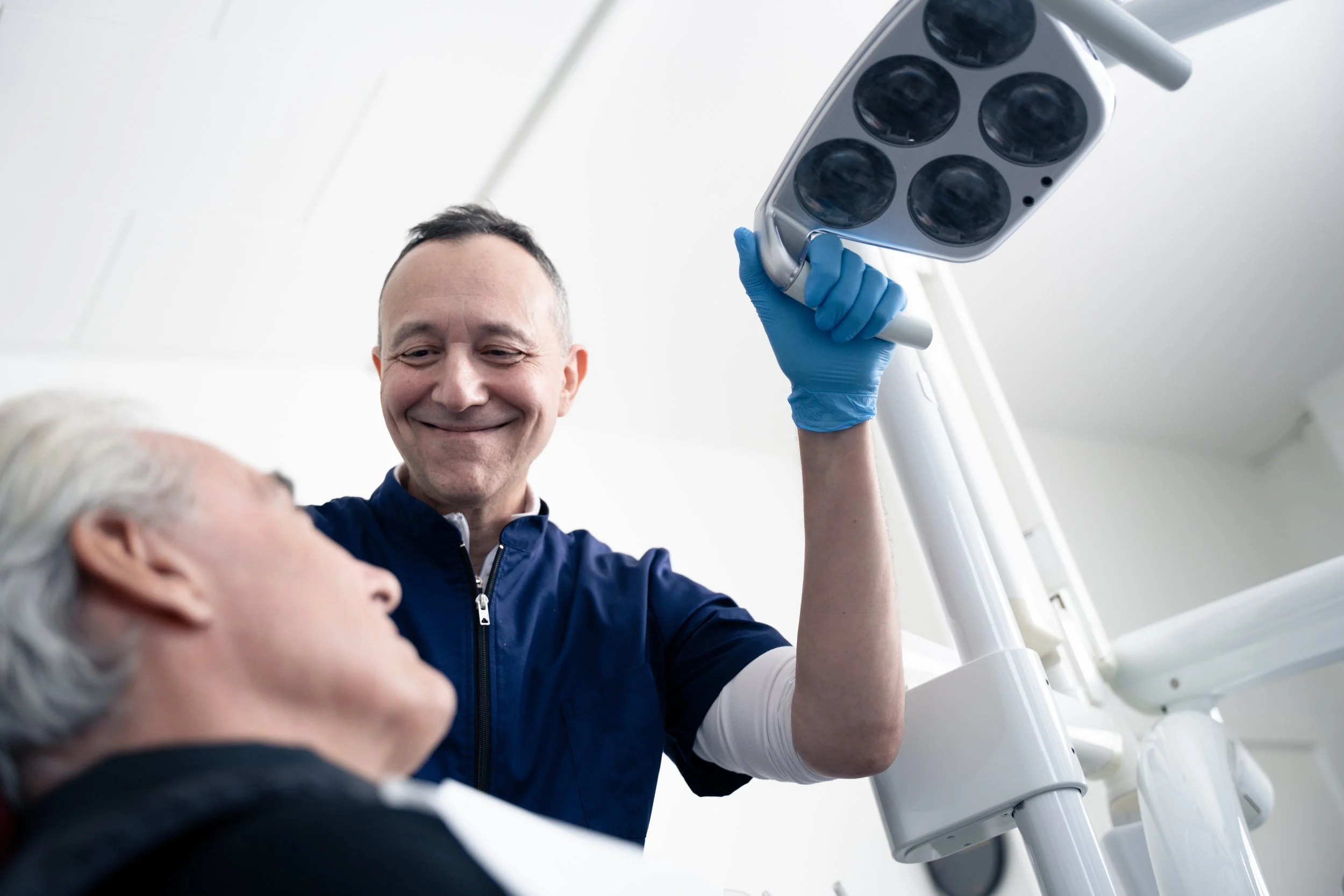 En mand i medicinsk uniform står ved siden af en syg patient på hospitalsseng, holder en kirurgisk lampe op og smiler venligt.