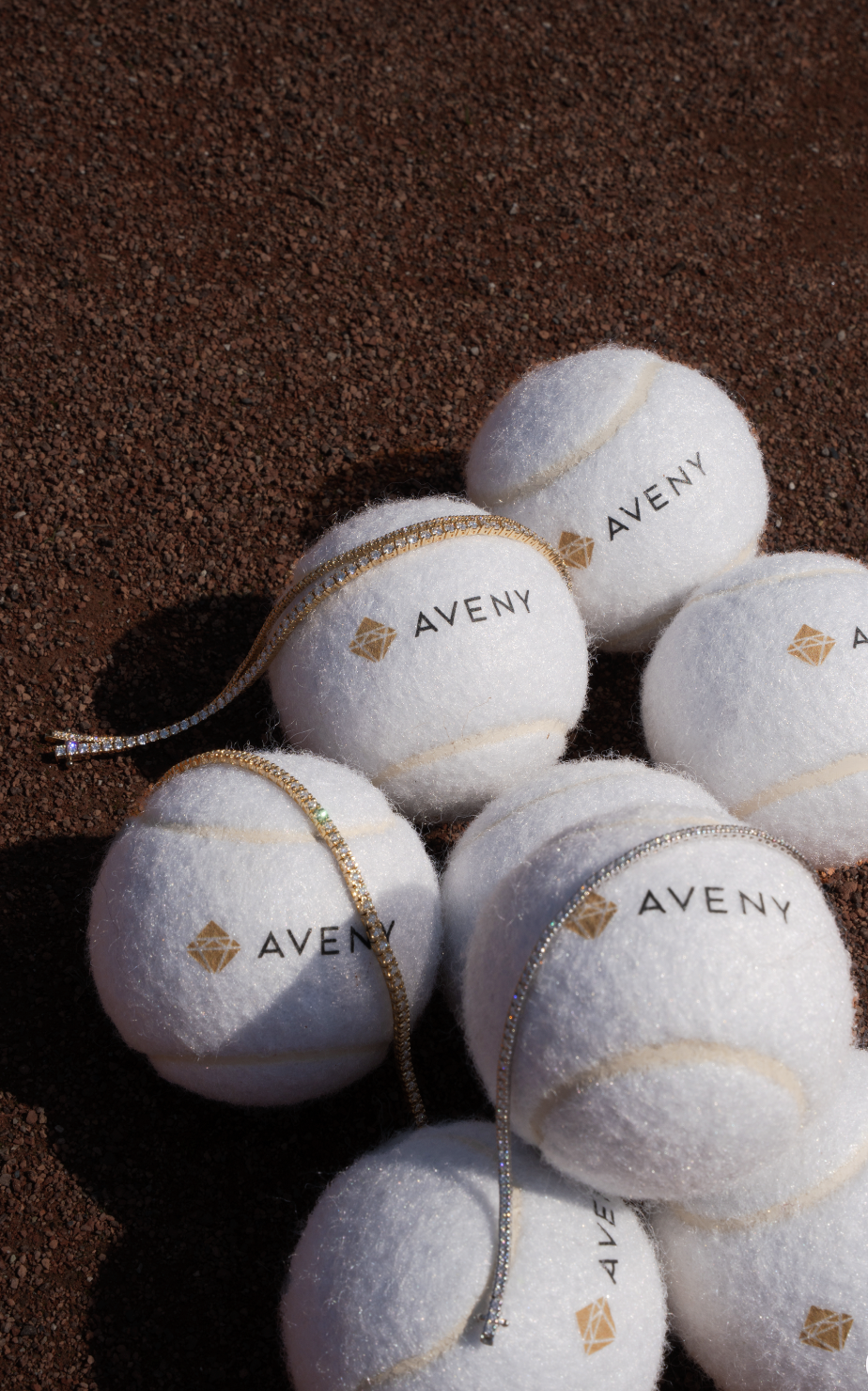 Hvide tennisbolde med logo og tekst 'AVENY' og smykker liggende på jord.