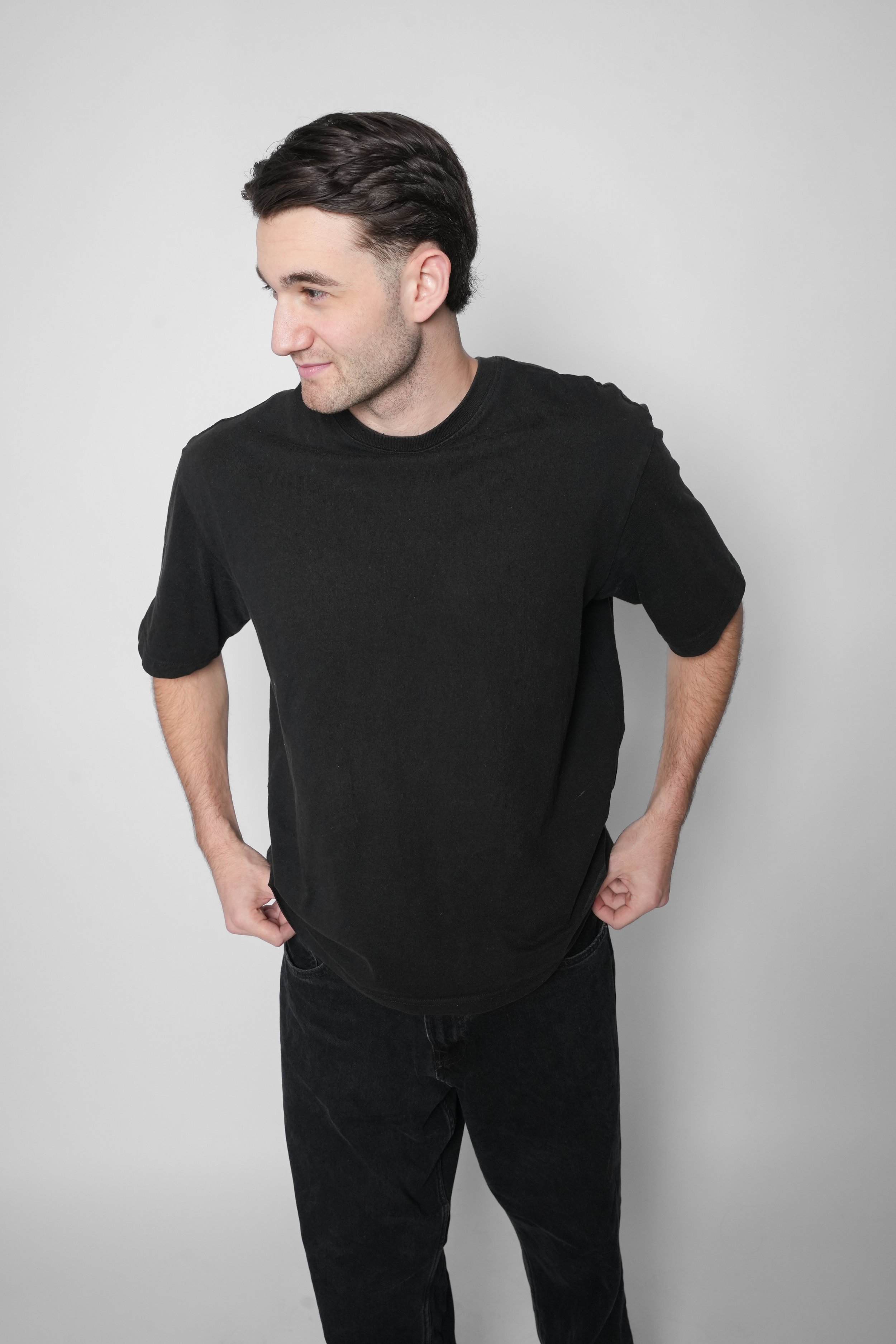 Ung mand i sort T-shirt og sorte bukser står mod en lys baggrund, set lidt i profil.