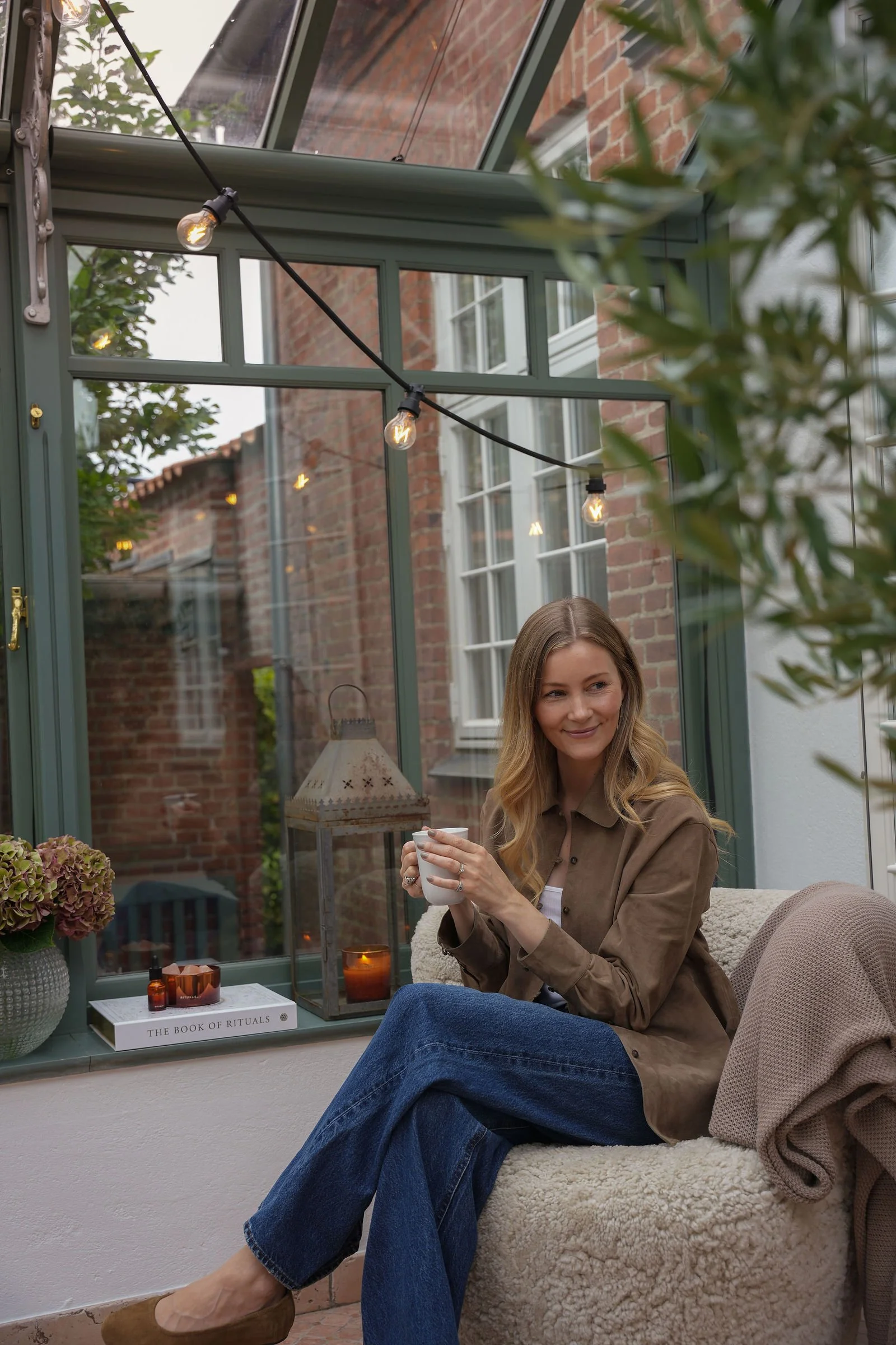 Kvinde sidder afslappet i et indendørs solrigt rum med store glasvinduer, holder en kop te, omgivet af stearinlys, planter og dekorative genstande som en bog og en lanterne.