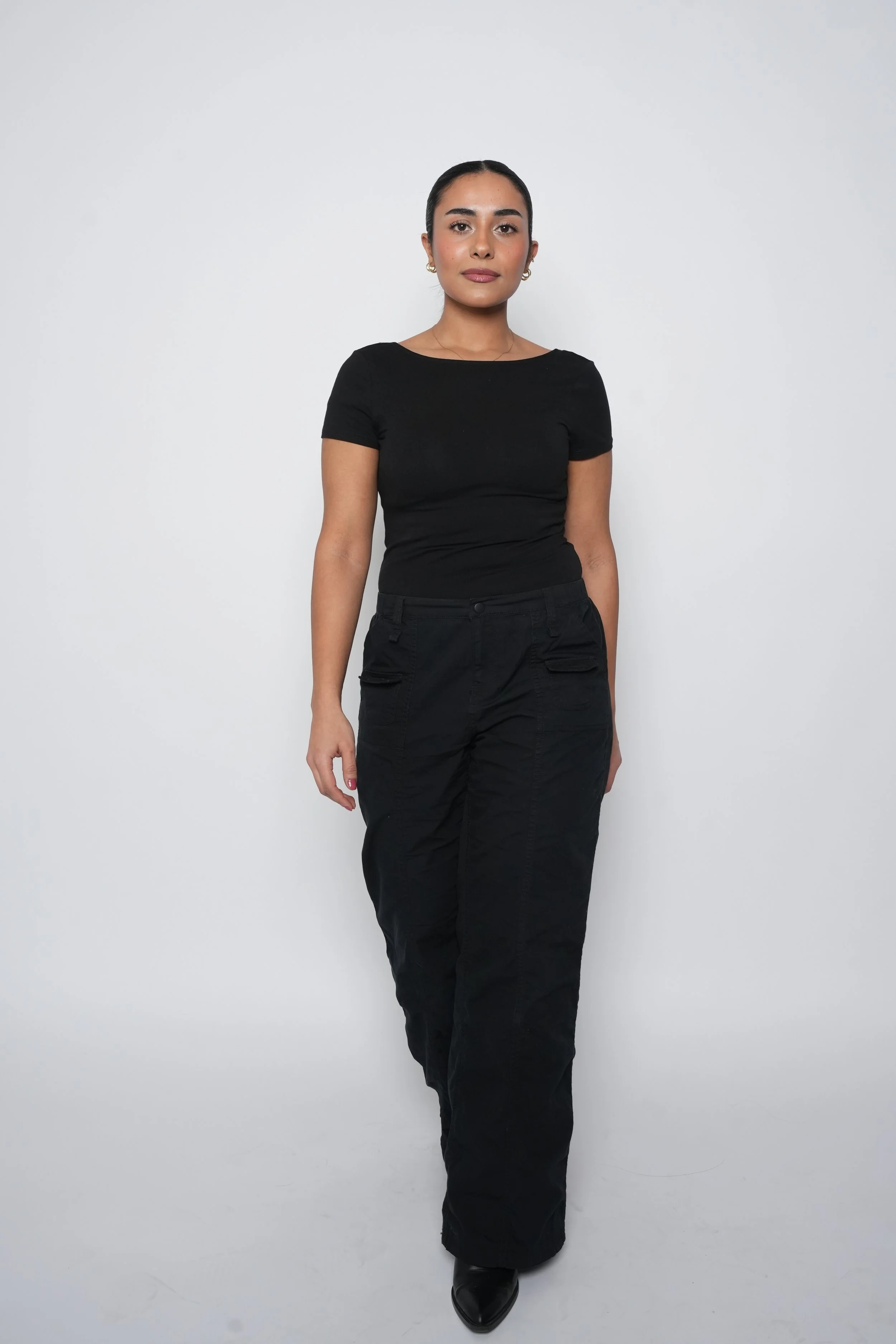 Kvinde går mod kamera med sort tøj og sorte sko, neutral baggrund.