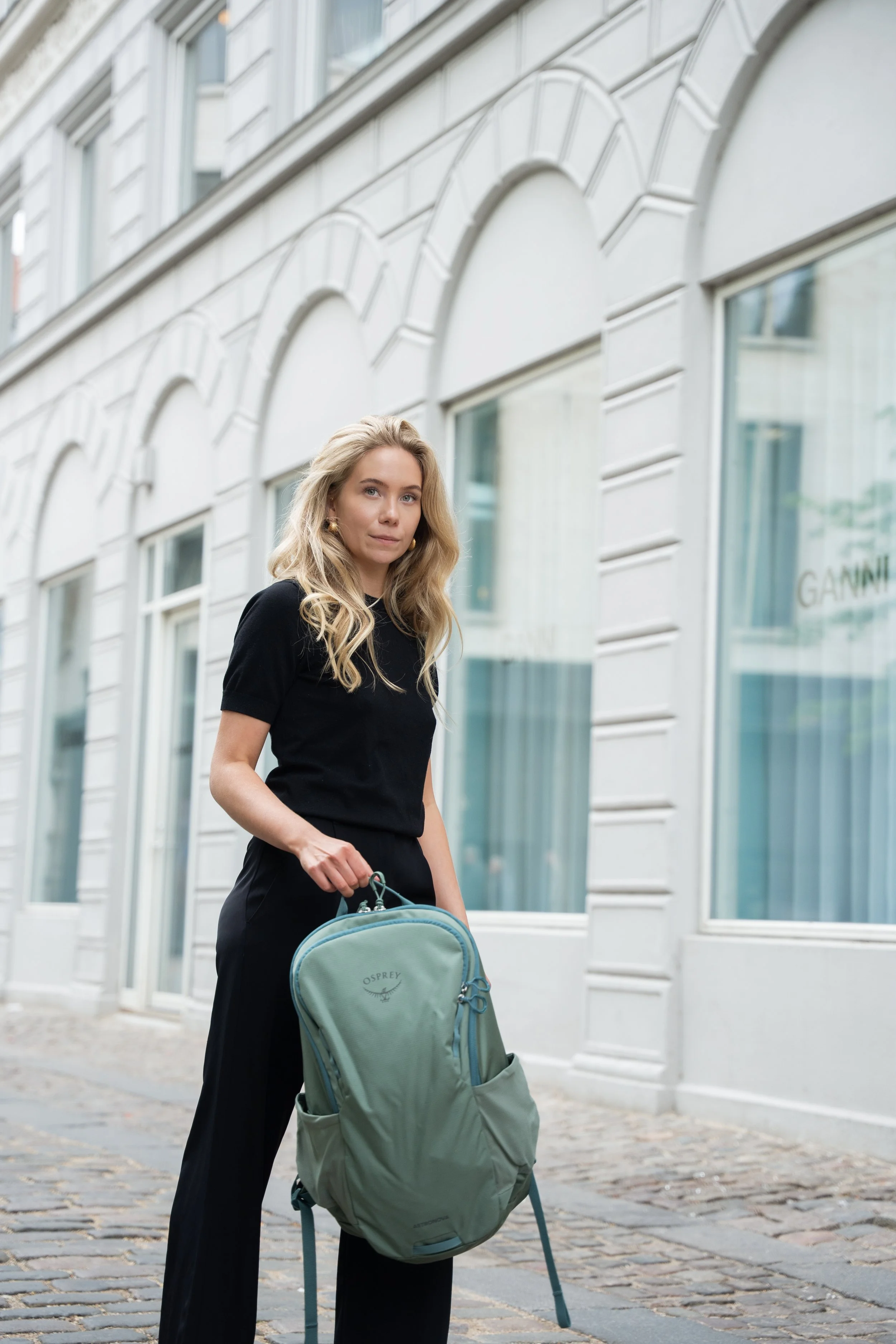 En ung kvinde med blond, bølget hår står på en brostensbelagt gade ud for en hvid bygning med store vinduer. Hun bærer en sort T-shirt, sorte bukser og holder en grøn Osprey rygsæk.