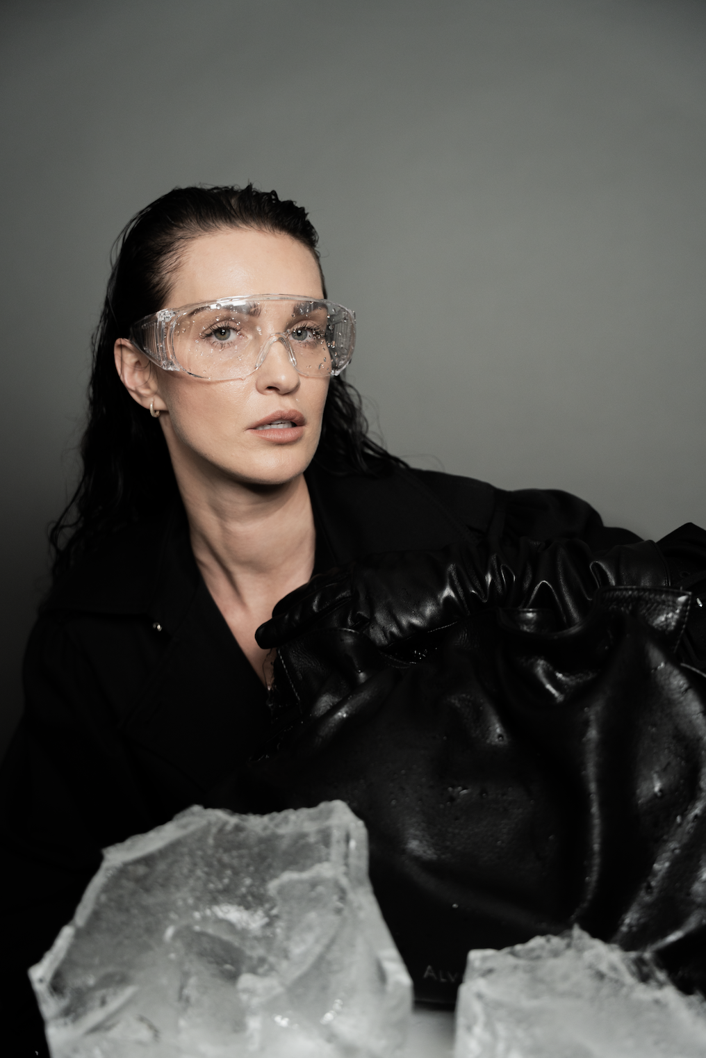 En kvinde med mørkt, viltert hår, iført beskyttelsesbriller og sort tøj, sidder foran knust is og lædertasker.