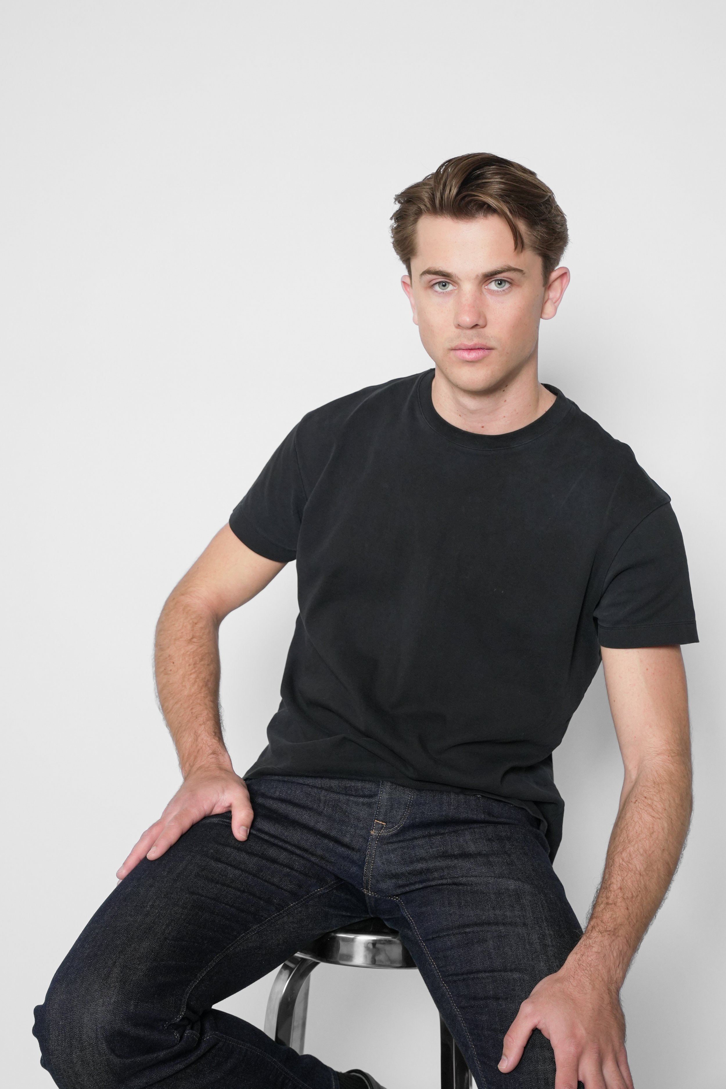 En ung mand med brunt hår, iført sort t-shirt og mørke jeans, sidder på en sølvfarvet stol mod en hvid baggrund.