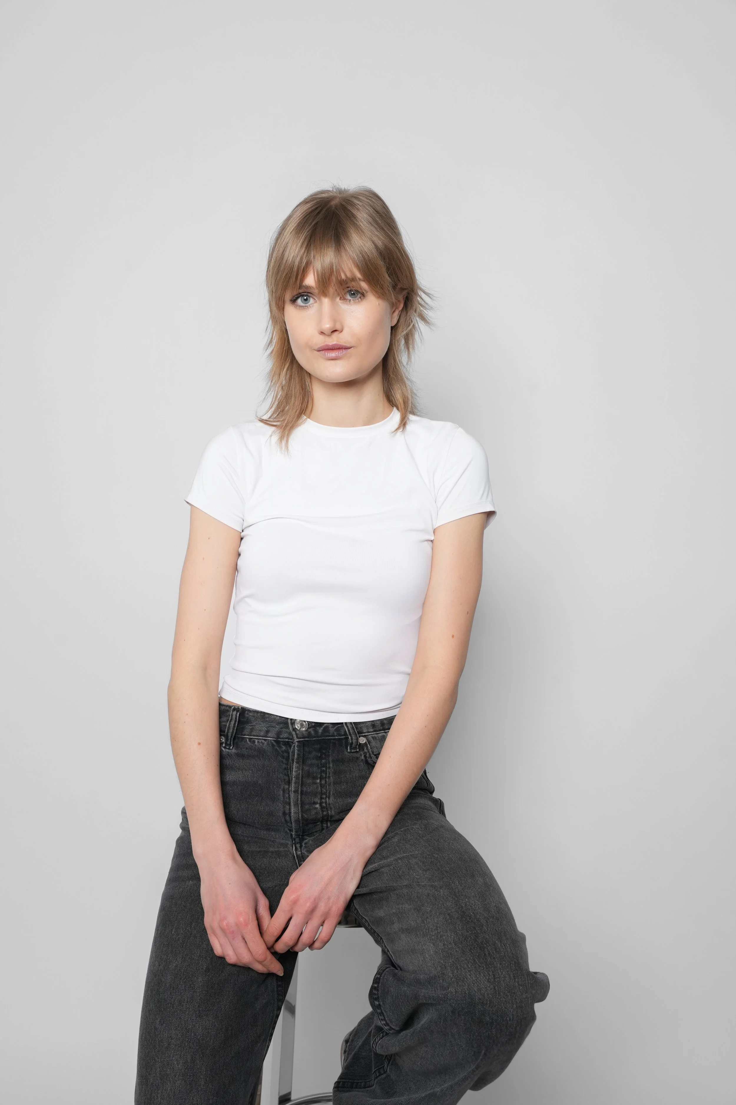 En ung kvinde med skulderlangt lysebrunt hår, iført en hvid T-shirt og mørke bukser, sidder på en barstol mod en hvid baggrund.