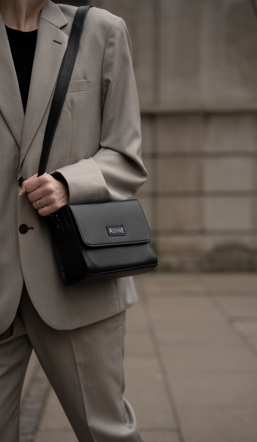 Kvinde iført beige jakkesæt, bærer en sort taske med mærket "ALVINÉ", over skulderen med en hånd, står foran en stenmur.