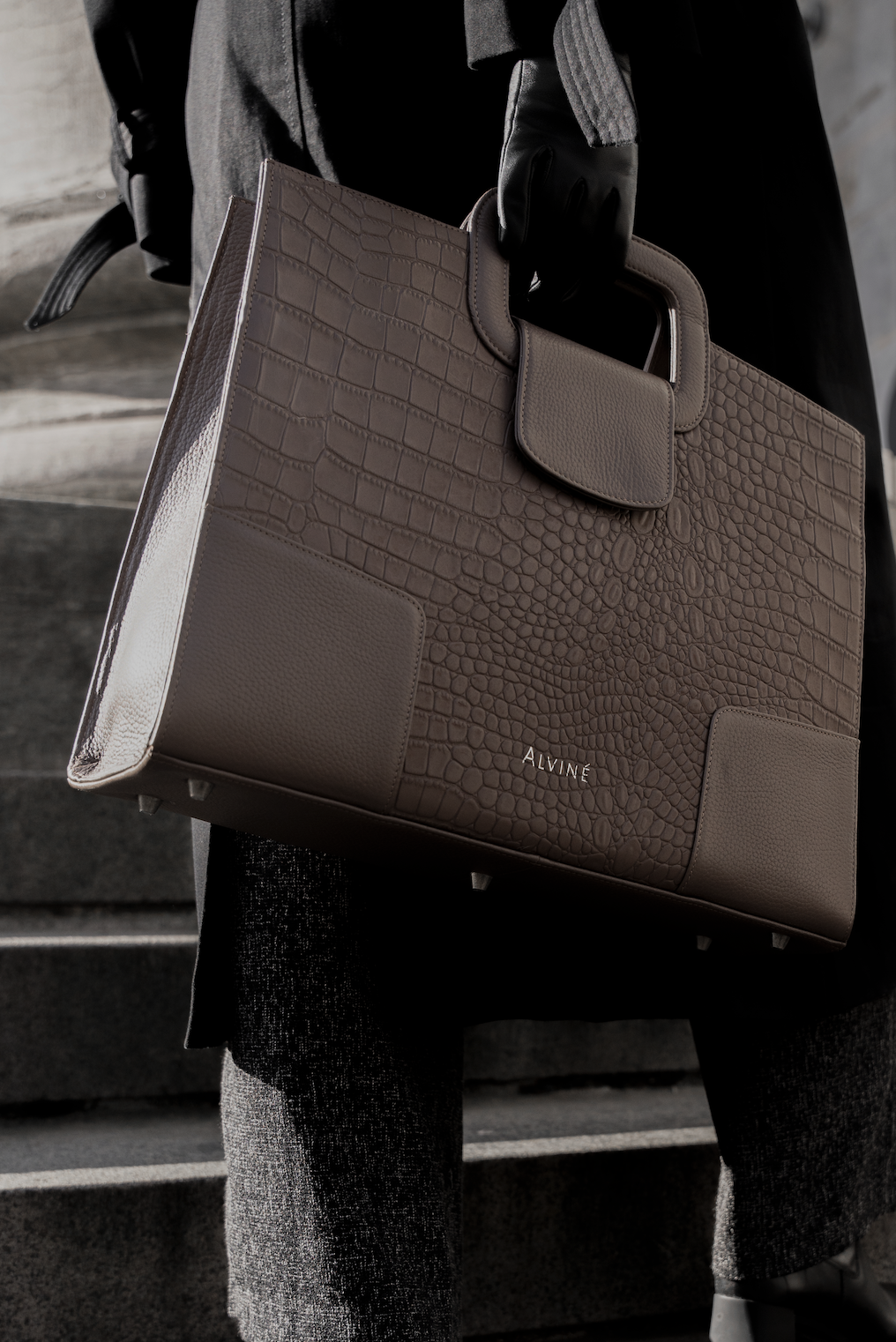 Person holder en stor taske i brune nuancer med crocodile-mønster og logoet ALVINÉ, mens de står på trappe.