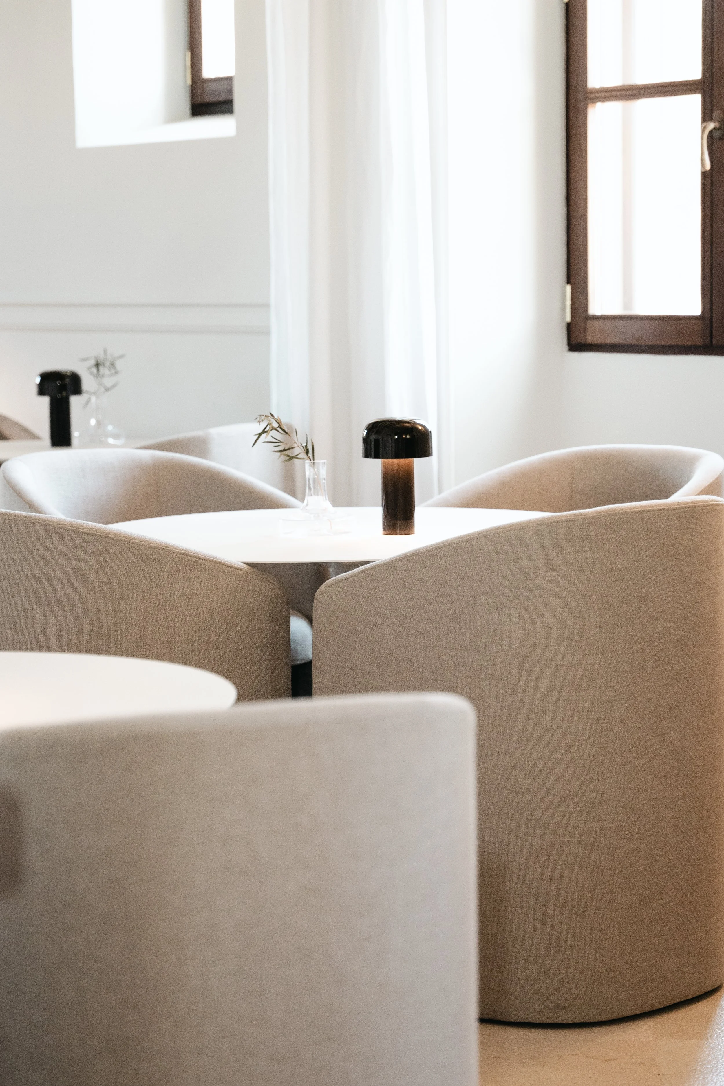 Et rundt bord med fire beige stole, dekorativt lys og en mindre vase med en gren, i et lyst rum med åbne vinduer og hvide gardiner.