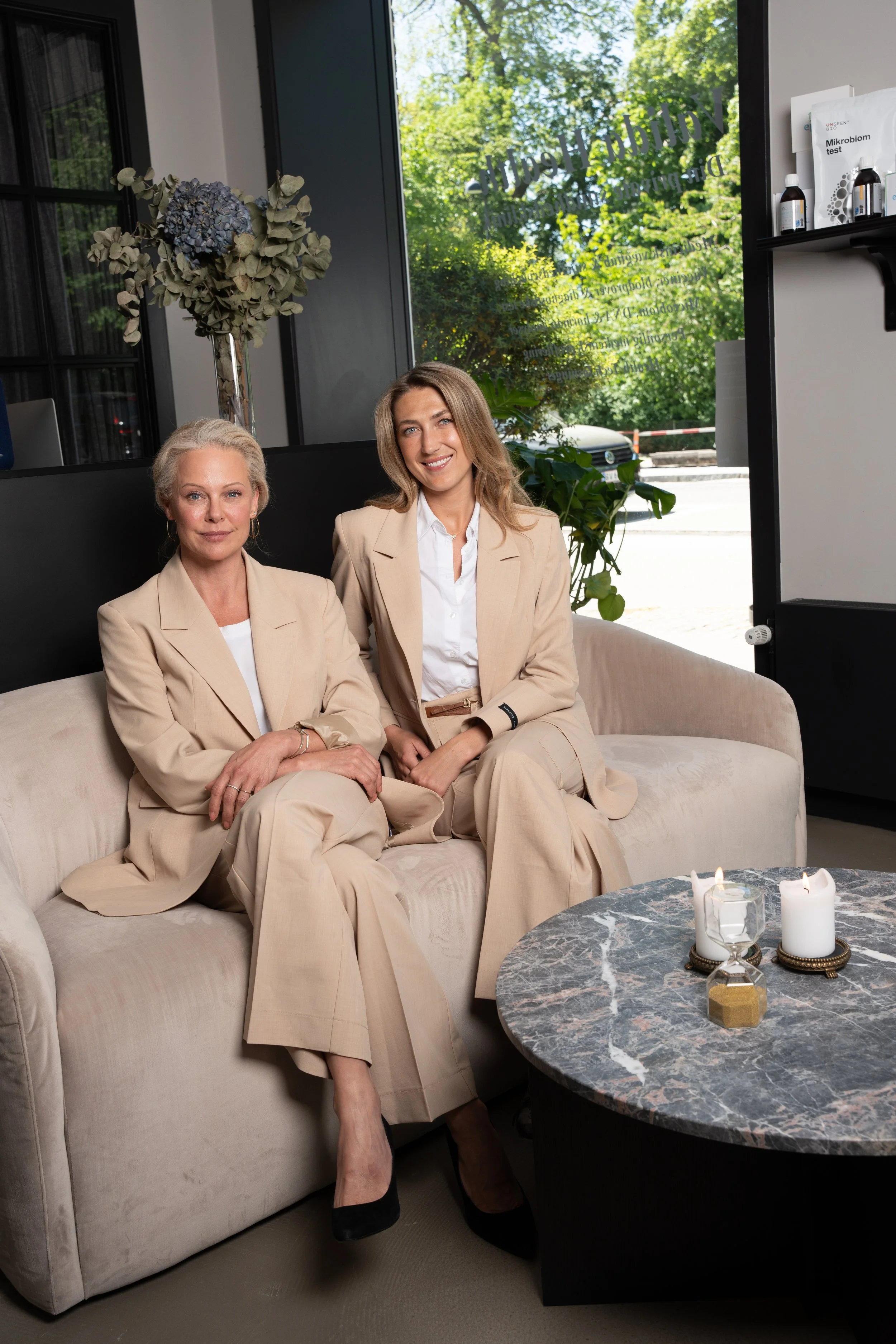 To kvinder sidder på en beige sofa i en moderne indretning, begge iført lyse beige jakkesæt. Der er et rundt sofabord med stearinlys foran dem, og store grønne planter udenfor vinduet bag dem.