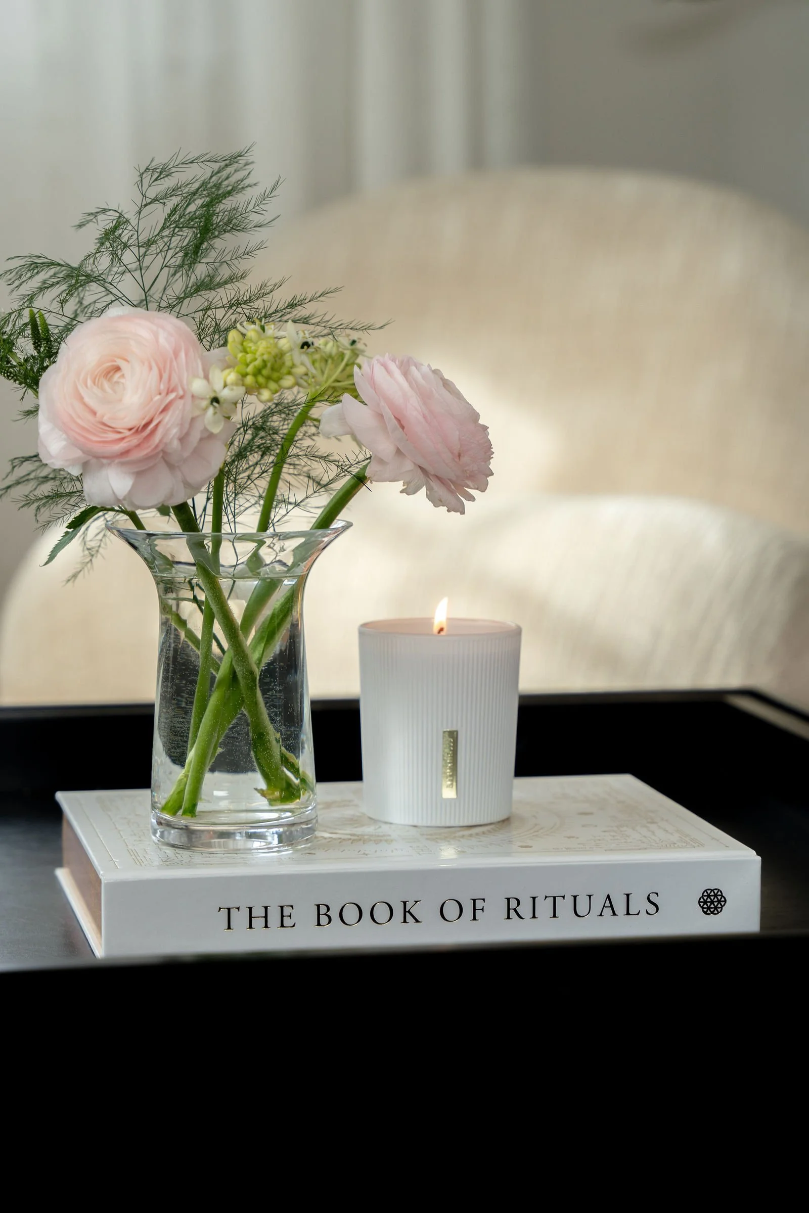 En vase med blomster, et lys, en bog med titlen 'The Book of Rituals' og en sætning på forsiden, placeret på en bog, sandsynligvis i et vindue.