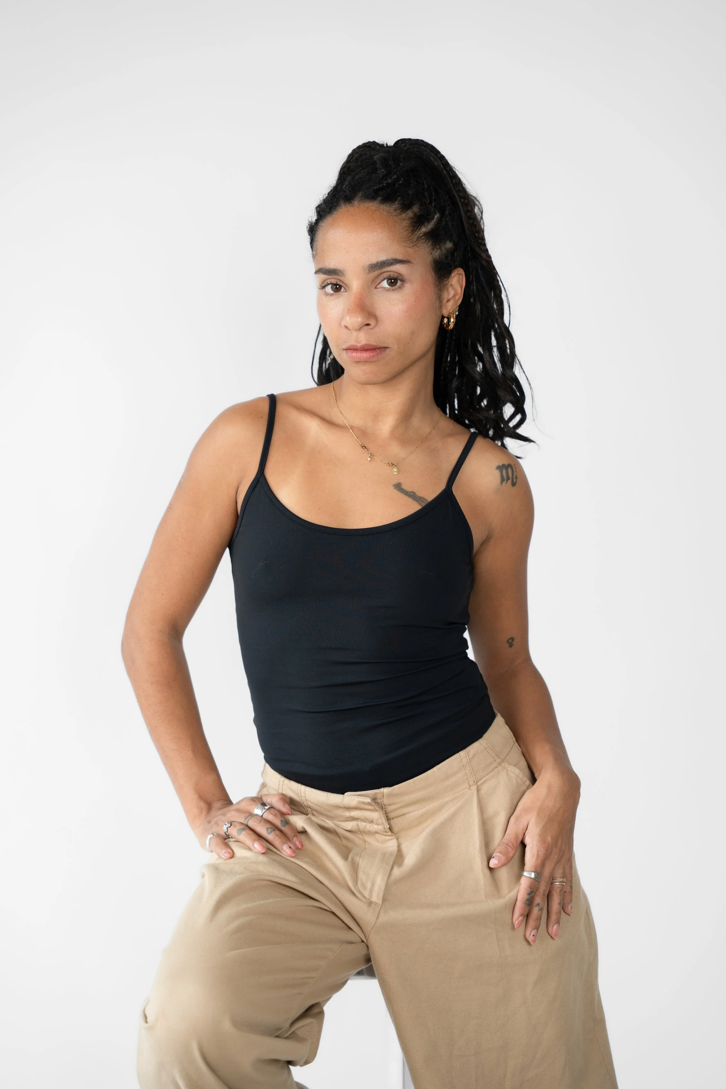 En ung kvinde med mørkt, krøllet hår i dreadlocks, iført sort tanktop og beige bukser, poser med hånden i lommen, står foran en hvid baggrund.