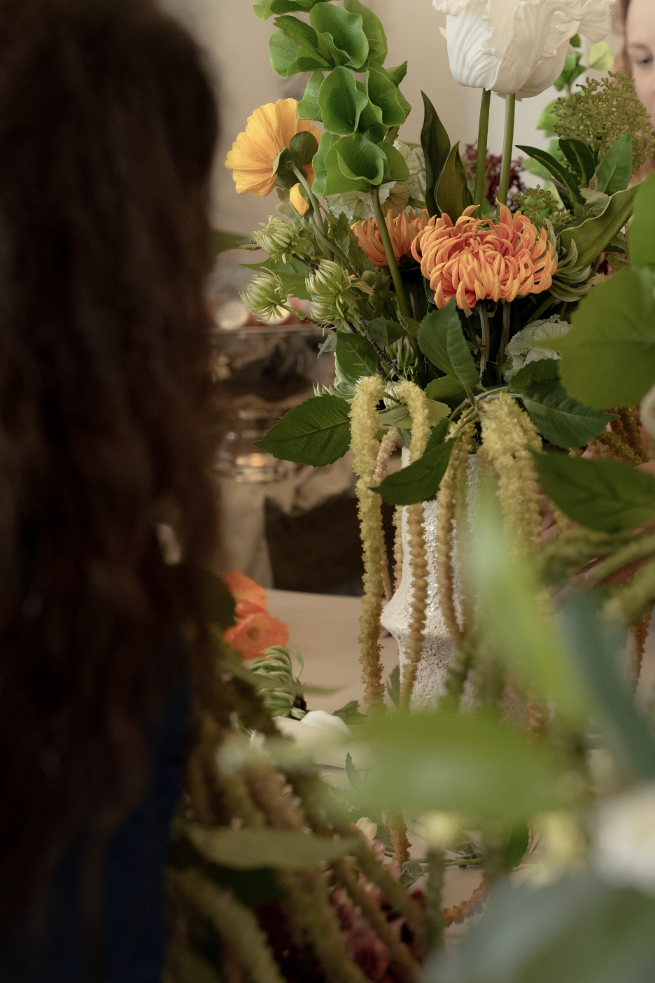En buket blomster med orange, hvid og grønne blomster og blade, set delvist bagfra.