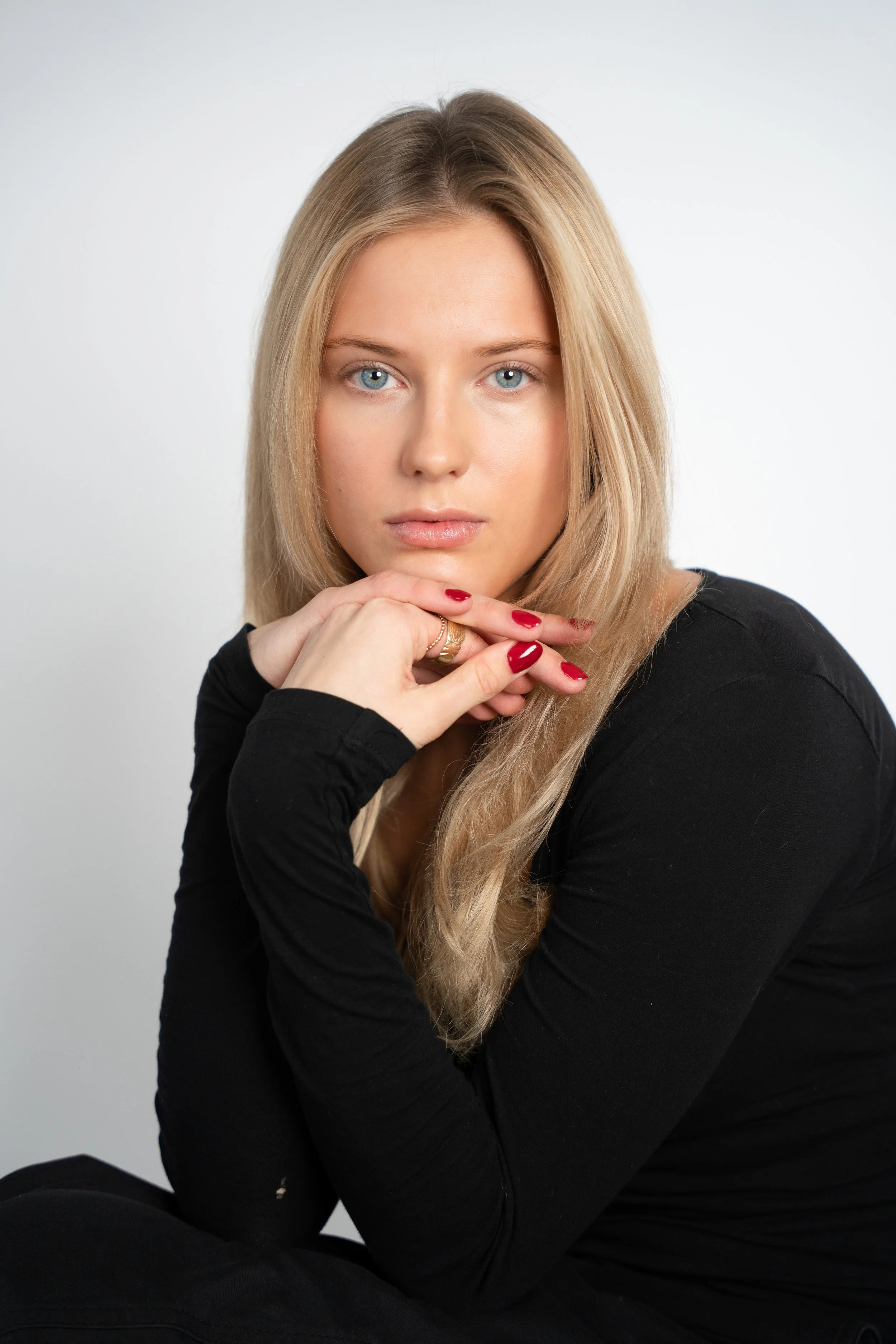 Portræt af en ung kvinde med blondt hår, blå øjne og rød neglelak, iført sort top, mod en lys baggrund.