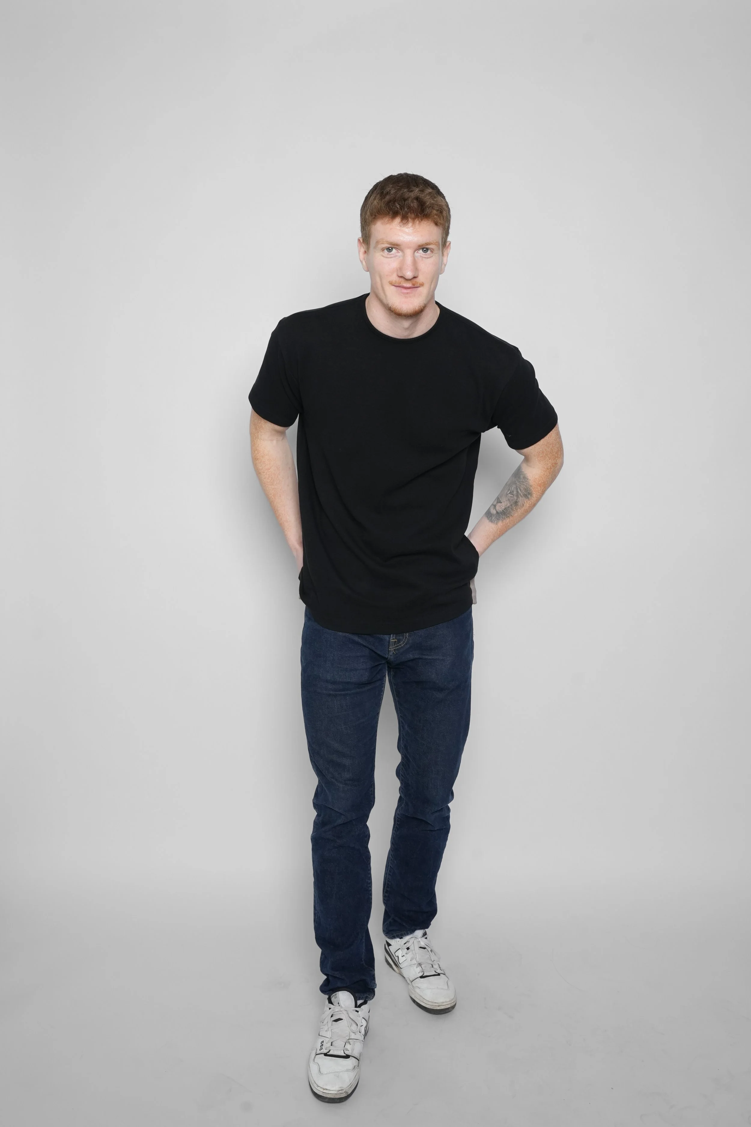 En ung mand iført sort t-shirt, jeans og sneakers står mod en lys grå baggrund og smiler let.