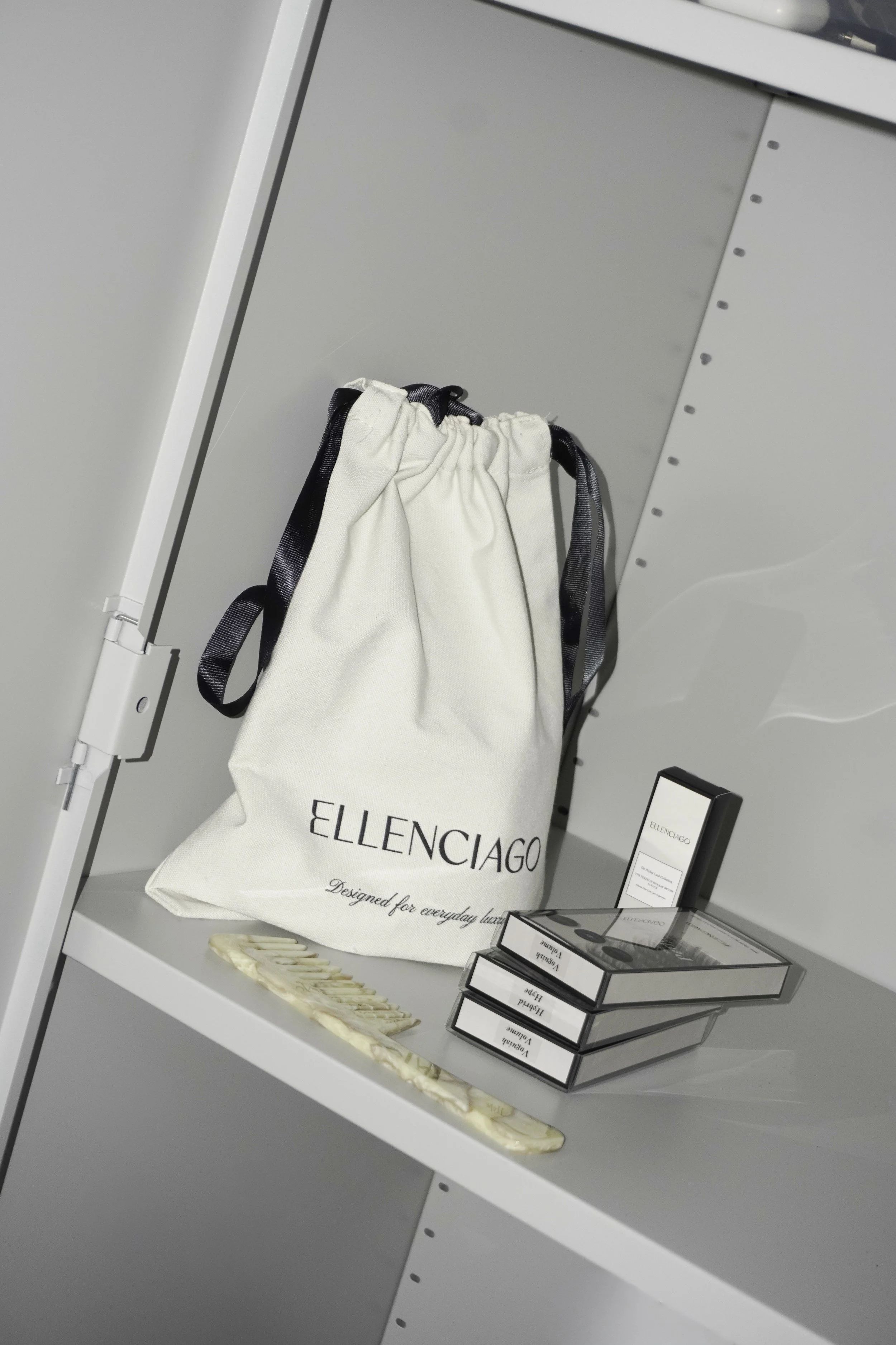 En hvid stofpose med teksten 'ELLENCIAGO' står på en hvid hylde, ved siden af er der tre sorte æsker, og en små, delvist synlig pakke står bagved. 