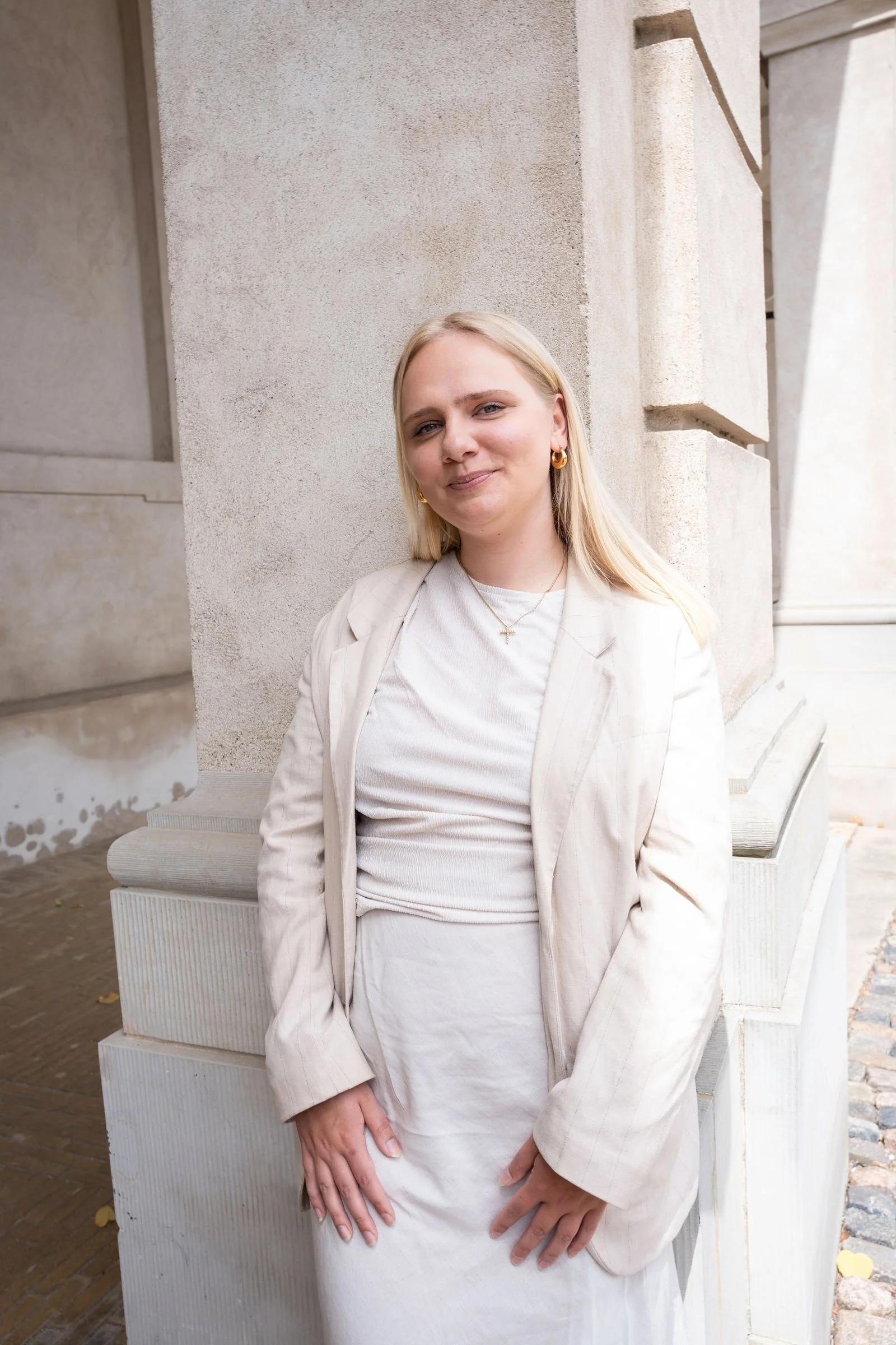 En smilende blond kvinde i hvidt jakkesæt står foran en lys stenvæg udendørs.