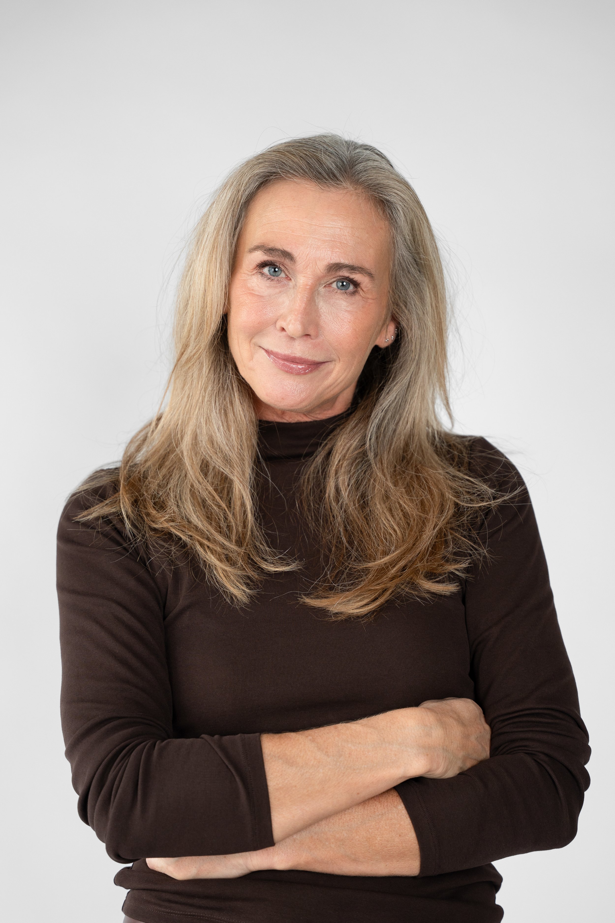 En midaldrende kvinde med langt, blond hår, iført en sort langærmet trøje, smiler let, står med foldede arme mod en neutral hvid baggrund.