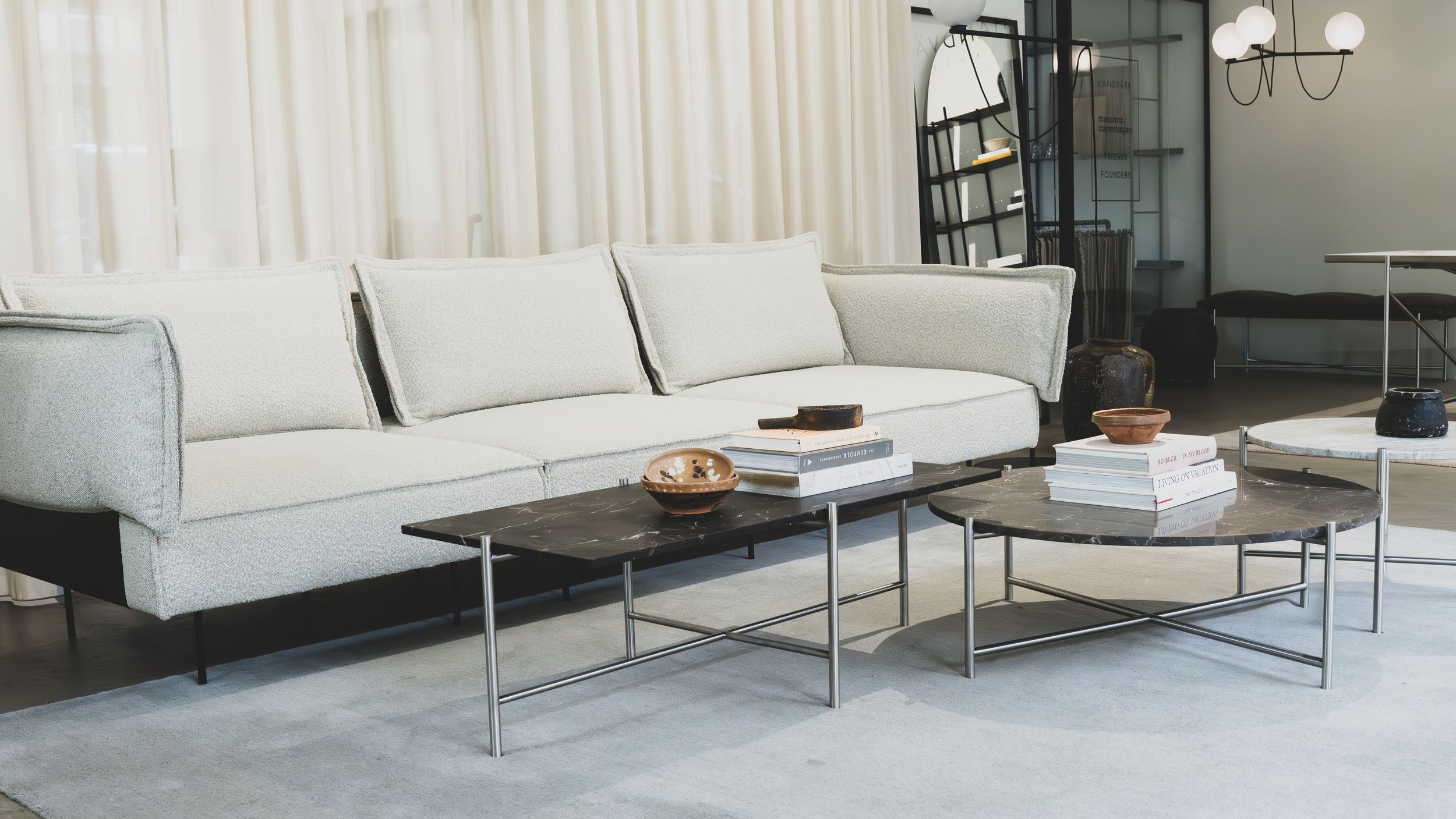 Moderne stue med hvid sofa, to sort marmorborde med bøger og dekorative genstande, og en baggrund af store vinduer med hvide gardiner.