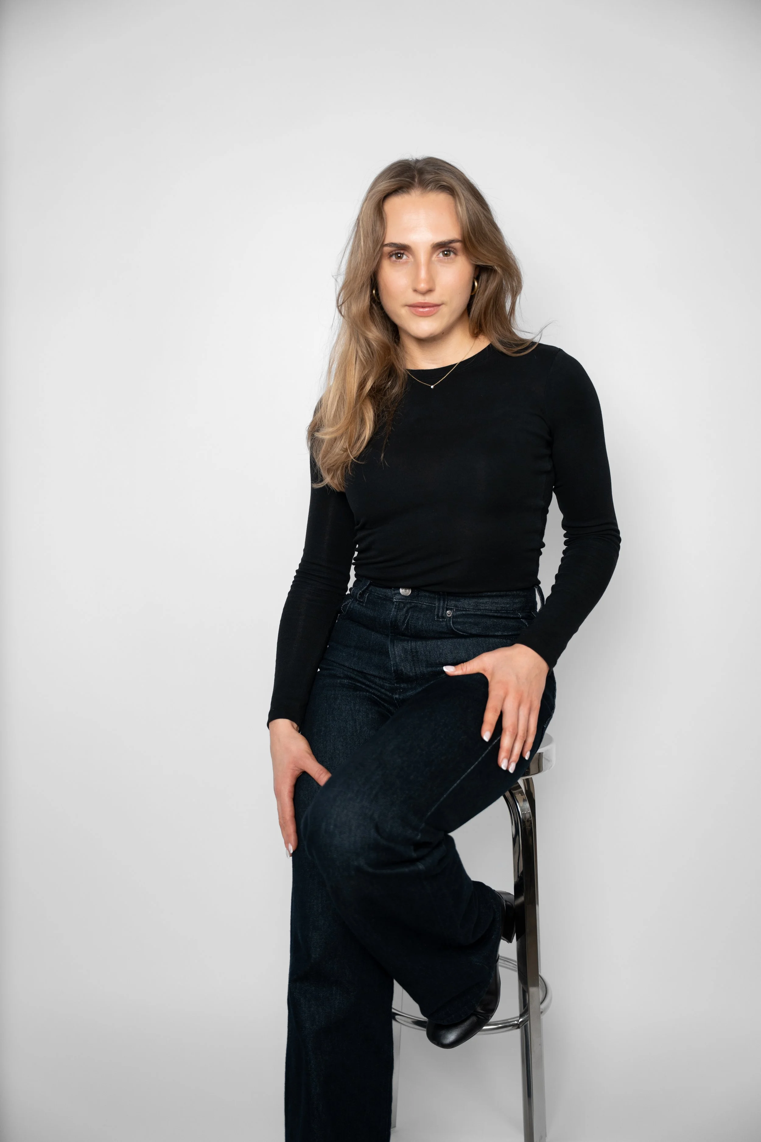 Kvinde sidder på en stol mod en hvid baggrund, iført sort langærmet top og mørke jeans.