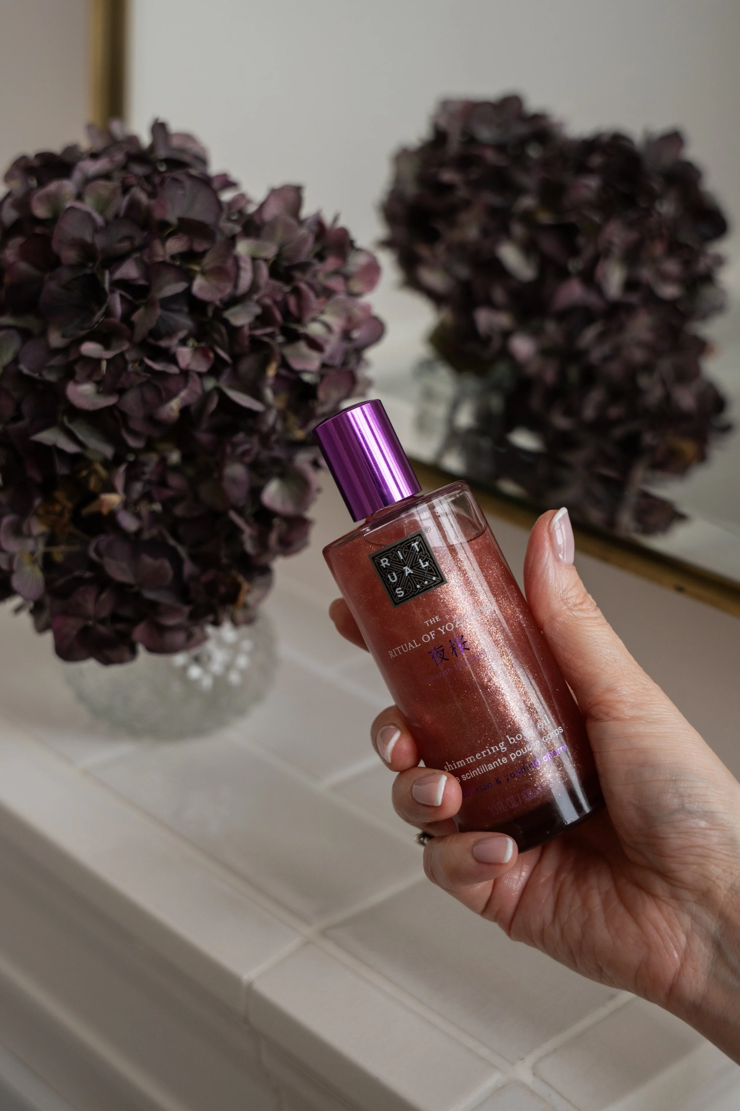 En hånd holder en flaske med shower gel, foran en vase med tørrede lilla blomster og et spejl.