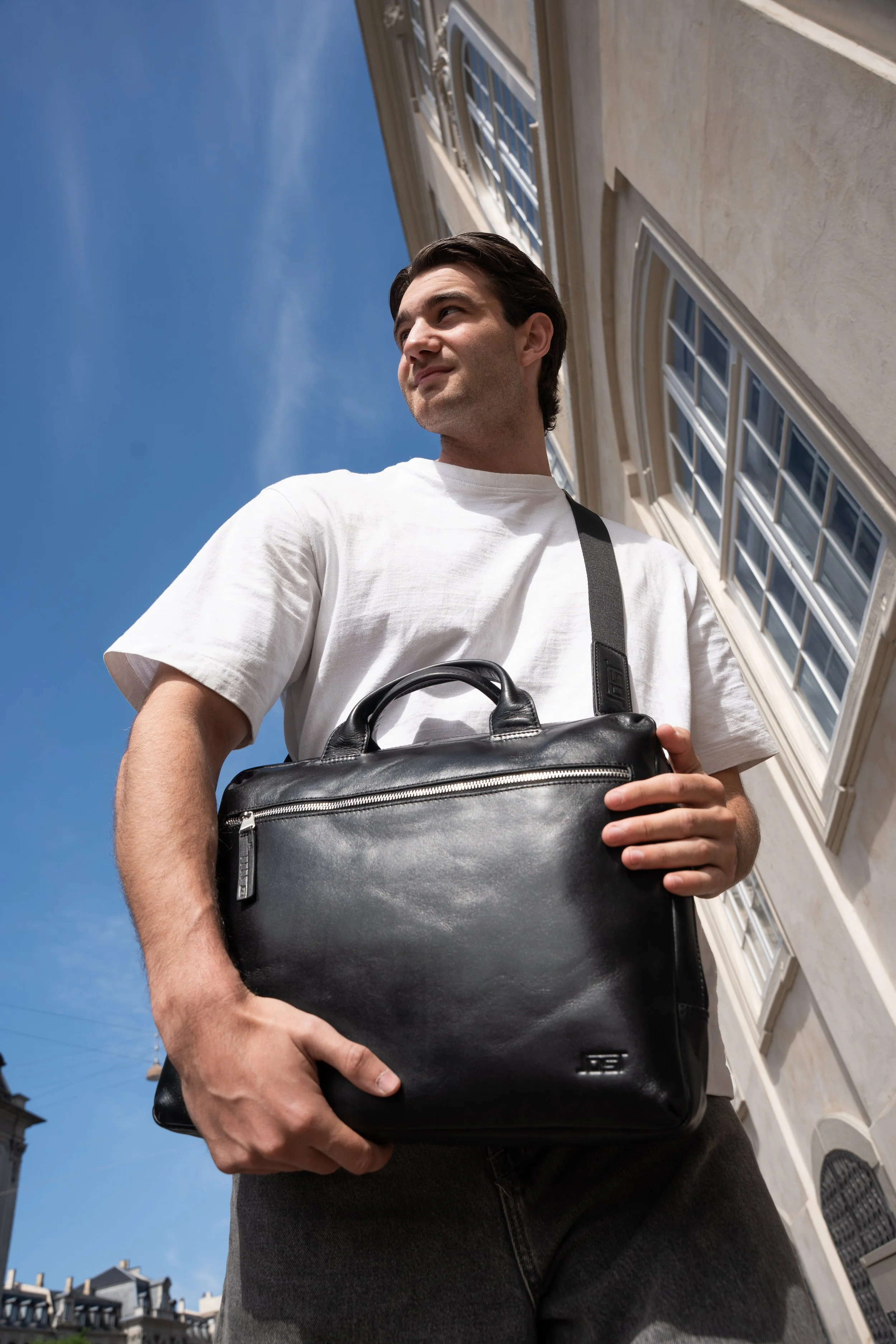 Mand, der går udendørs med en sort taske i hånden, set oppefra, foran et bygning med store vinduer og en blå himmel.