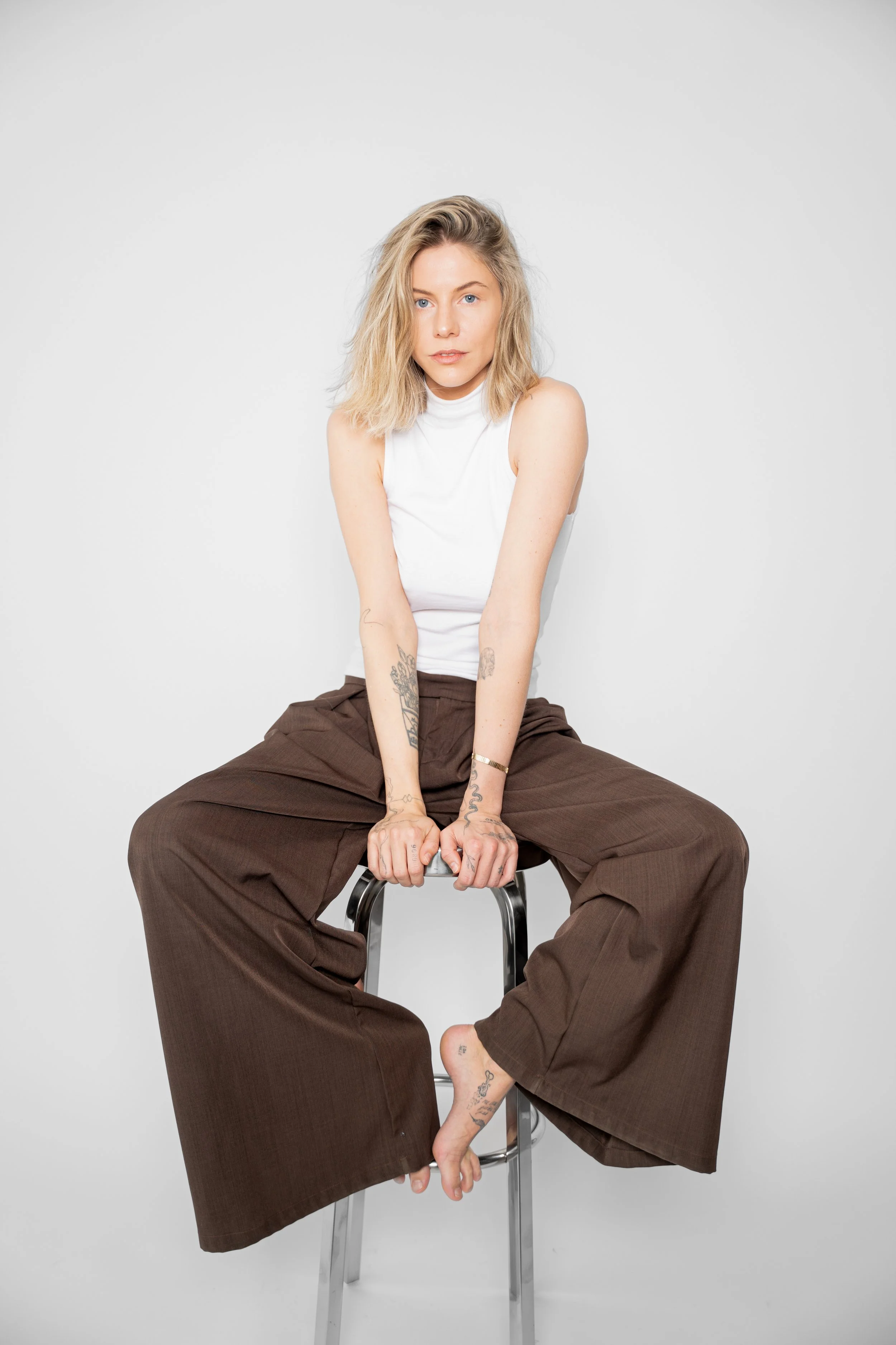 Kvinde sidder på en barstol mod en hvid baggrund, iført hvid top og brede brune bukser, med tatoveringer og blond hårlok.