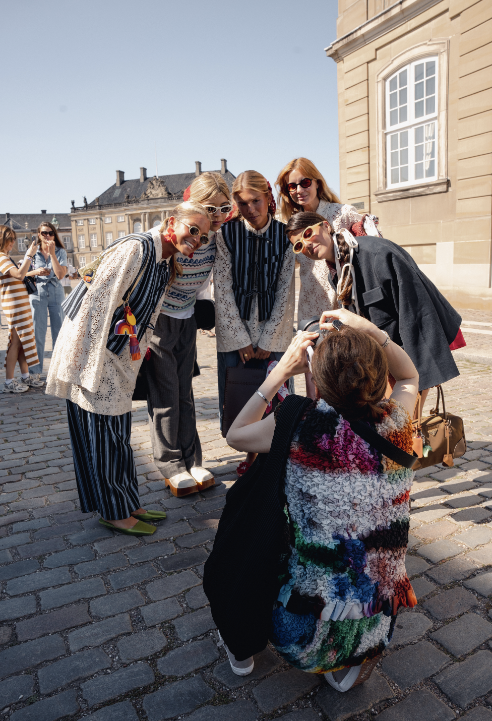 Fem kvinder poserer for et photograph på brosten foran historiske bygninger. De smiler, ser trendy ud og bruger solbriller. En person tager billedet, mens de andre holder en gruppe sammen.