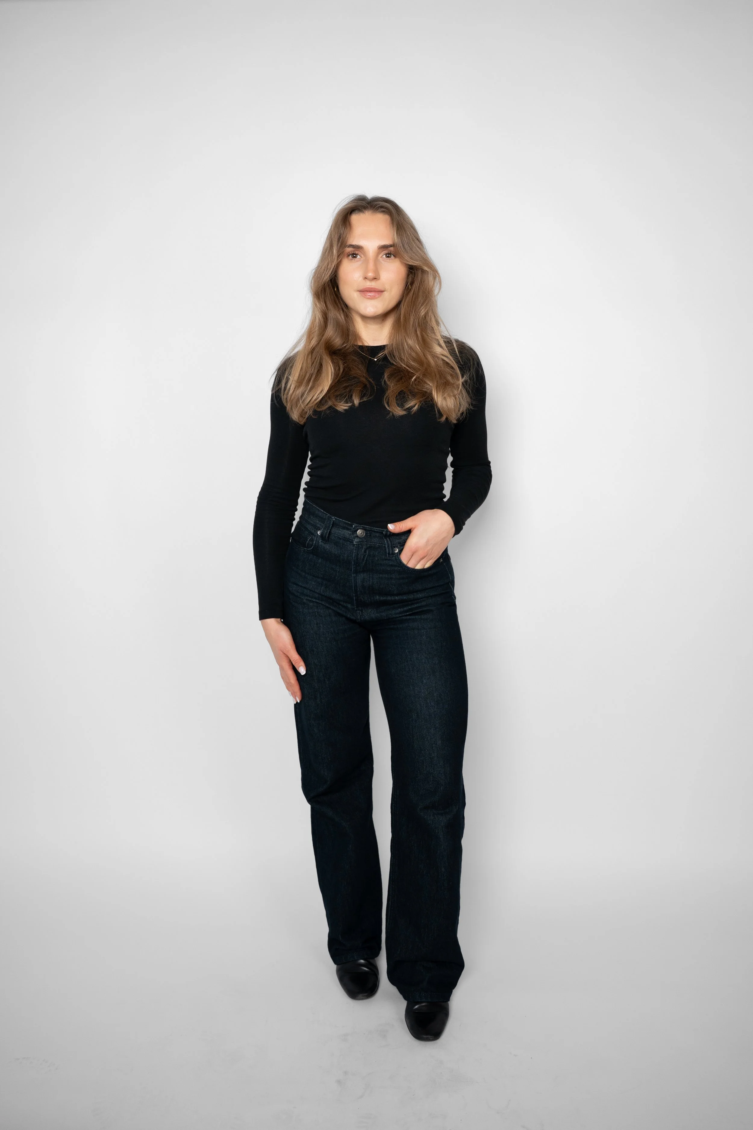 En ung kvinde med brunt, mellemlangt hår står foran en hvid baggrund, iført sort langærmet top og mørke jeans.