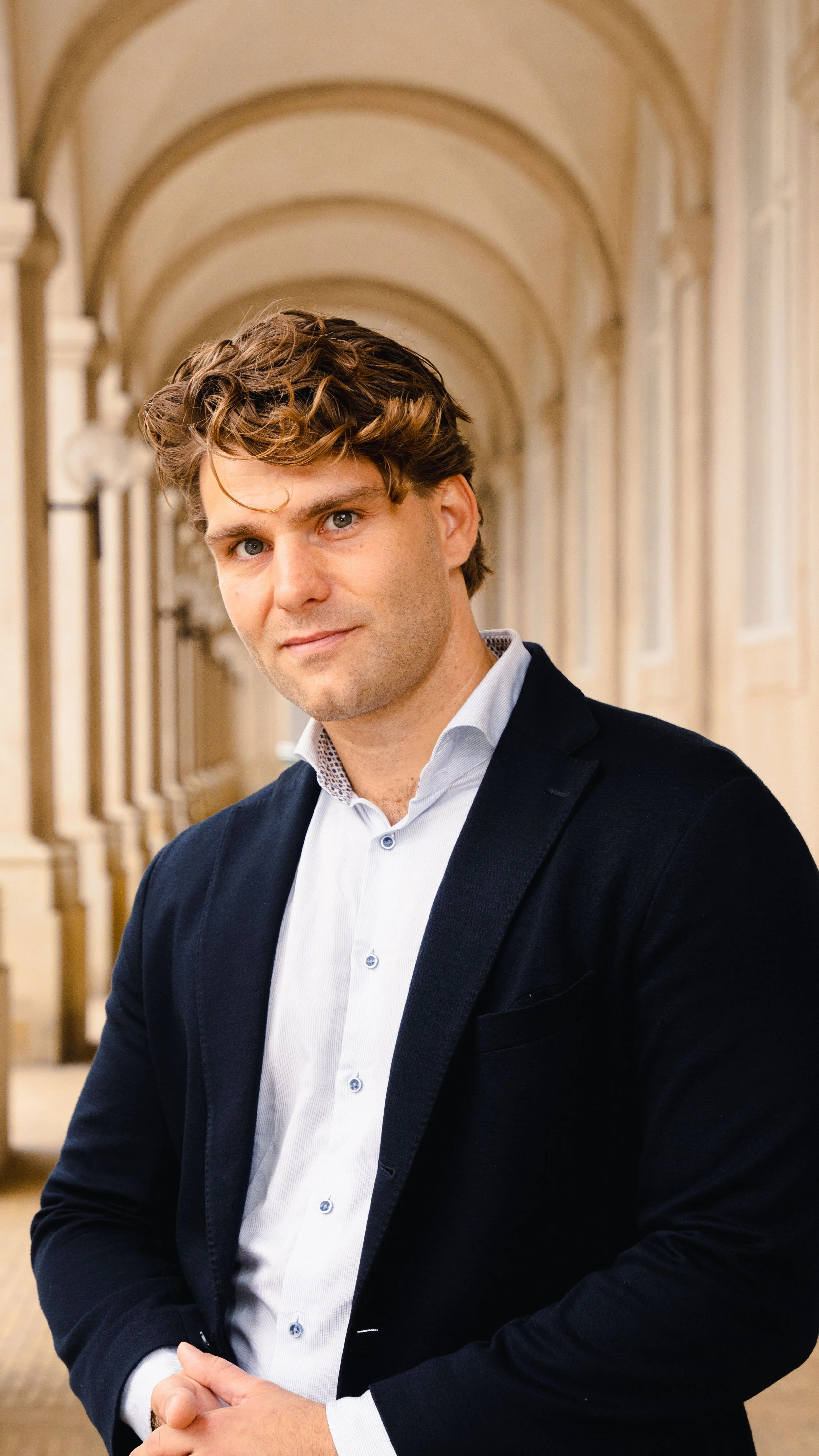 Portræt af en mand i formel tøj, stående i en arkitektonisk korridor med buede lofter og store vinduer.