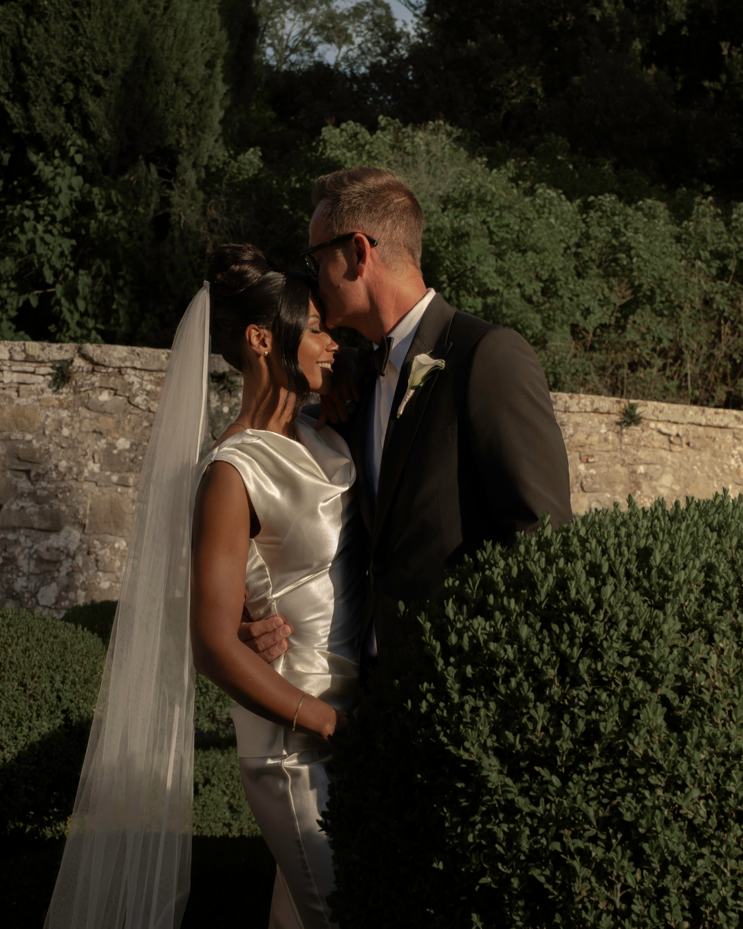 Et brud og en mand i brudekjole og smoking deler et kærligt øjeblik udenfor. De står bag en busk, omgivet af grønne planter og en stenmur.