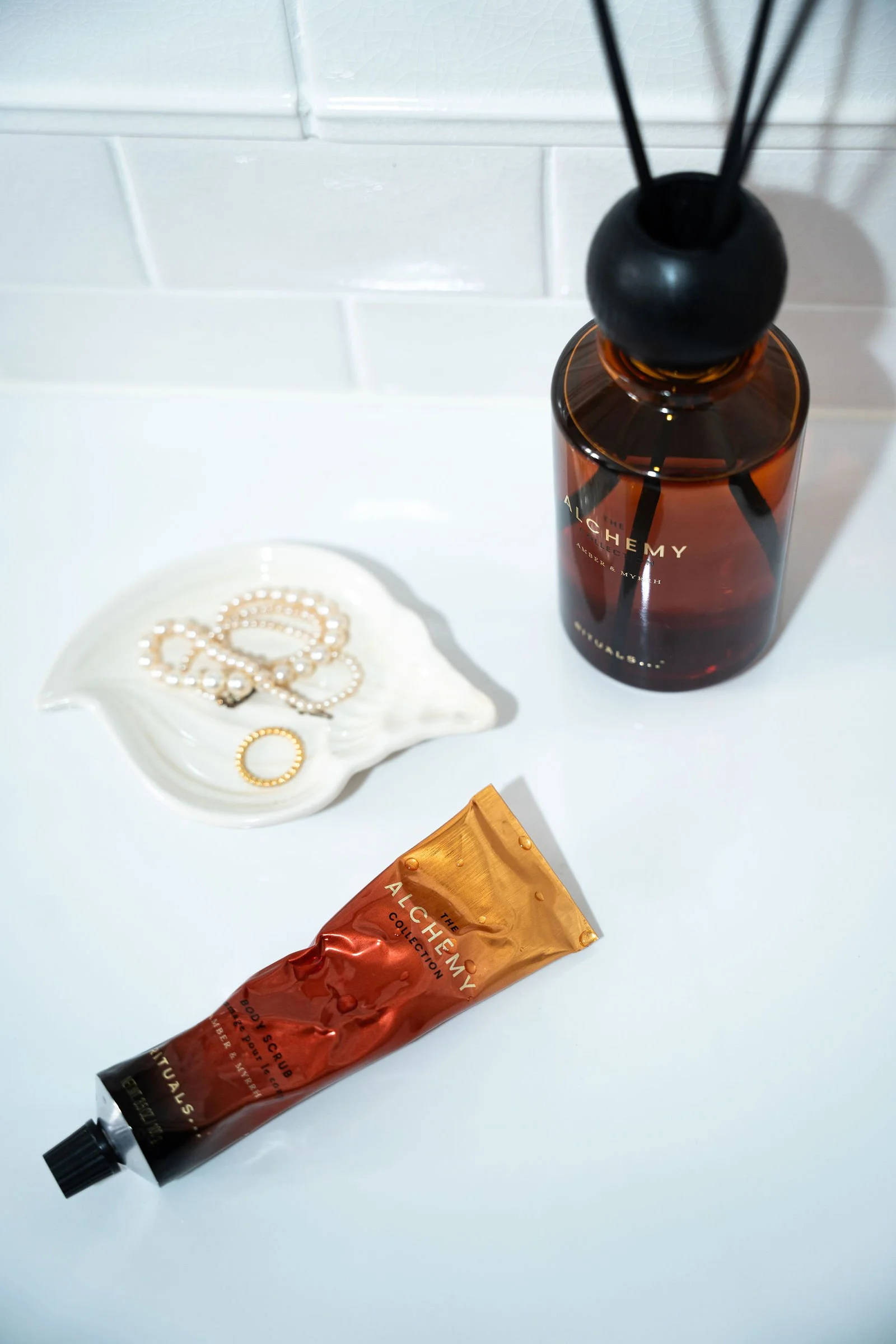 En flaske med duftolie, en tube med body scrub, en skål med perlekæder, og en sort diffuser med reed sticks.