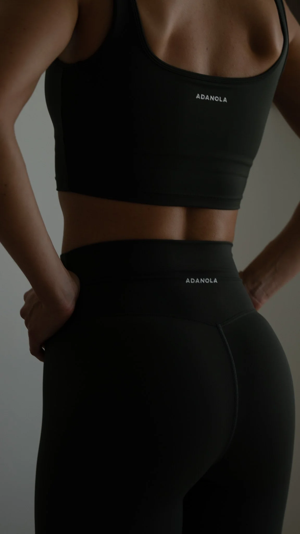 Nærbillede af en person i sort træningstøj, set bagfra, med fokus på midten af kroppen, der står med hænderne på hofterne.