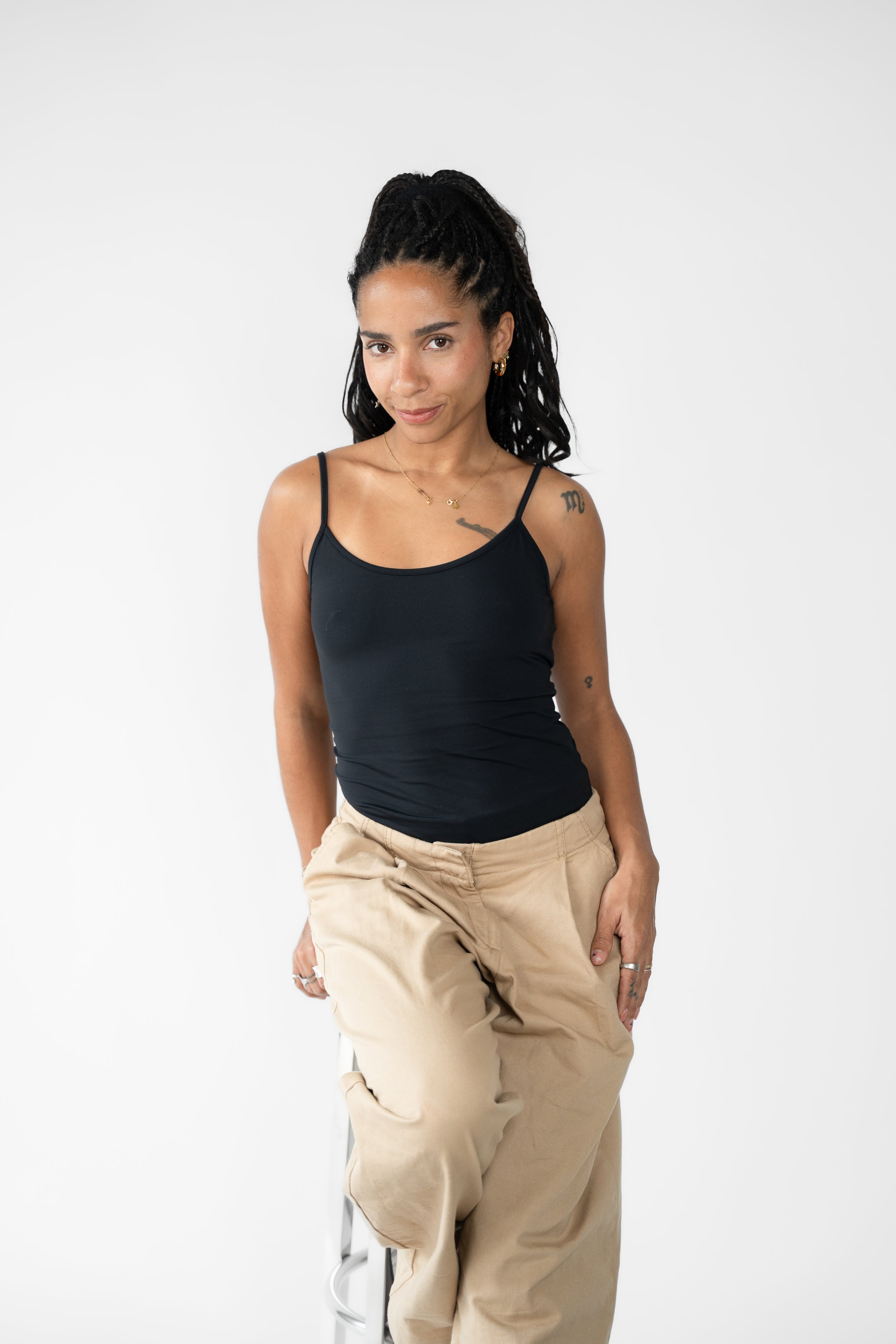 Kvinde med langt, krøllet hår, iført sort tanktop og beige bukser, sidder på en stol mod en hvid baggrund.