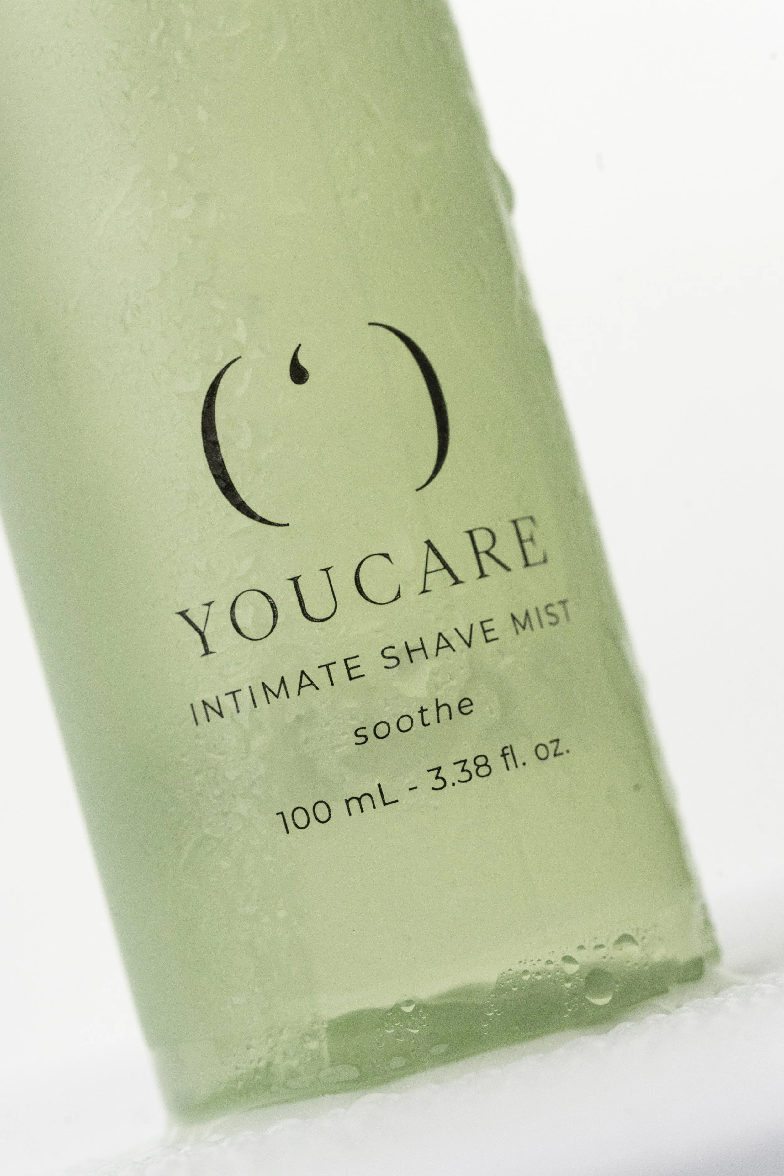 Et grønt sprayflaske med etikettet 'You Care Intimate Shave Mist', som indeholder 100 ml væske, og er dækket af vanddråber.