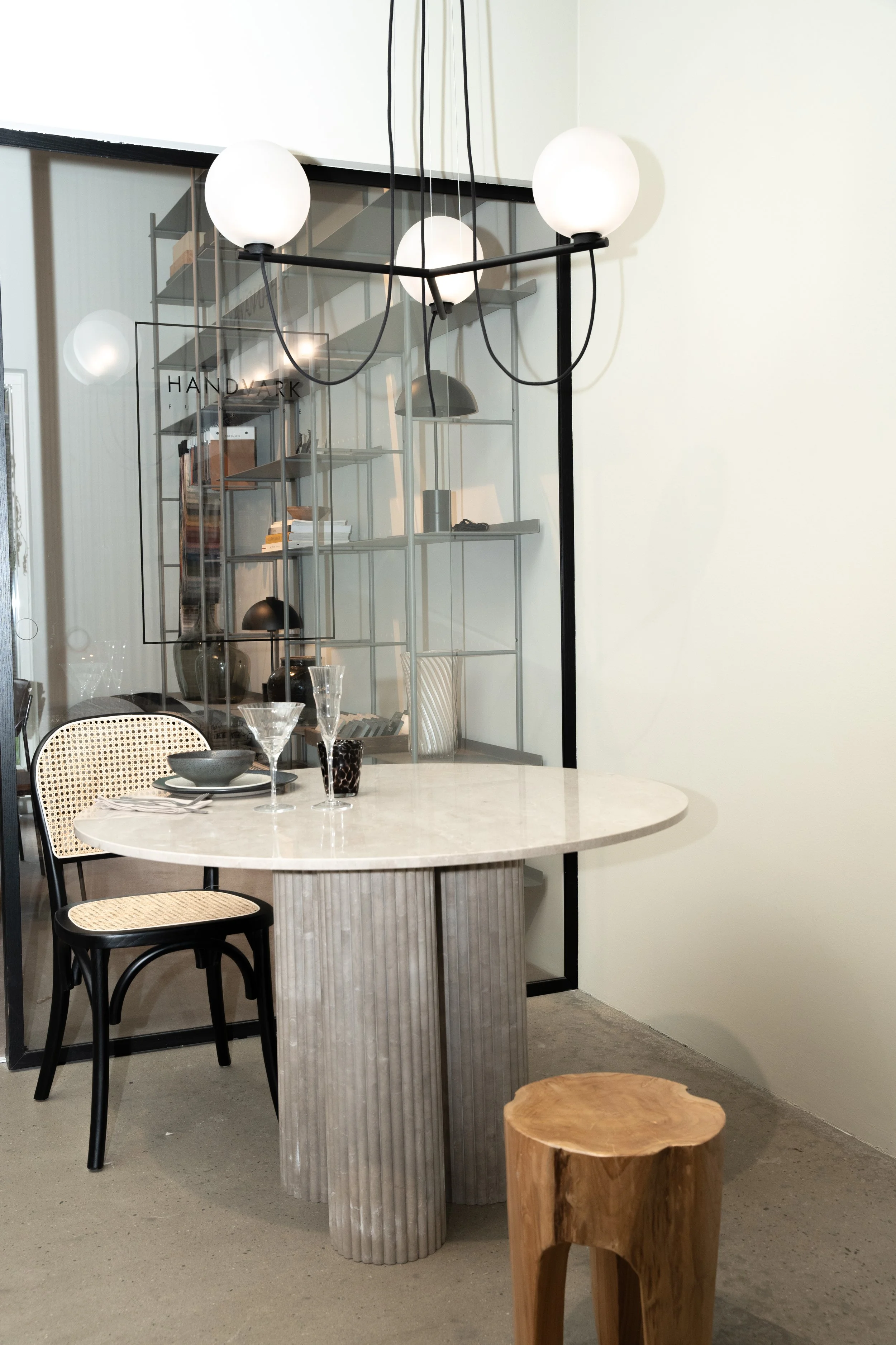 Et rundt bord med spiseudstyr, to stole og en lille træstol foran en væg, adskilt af en glasvæg med hylder og lamper, i en moderne indretning.