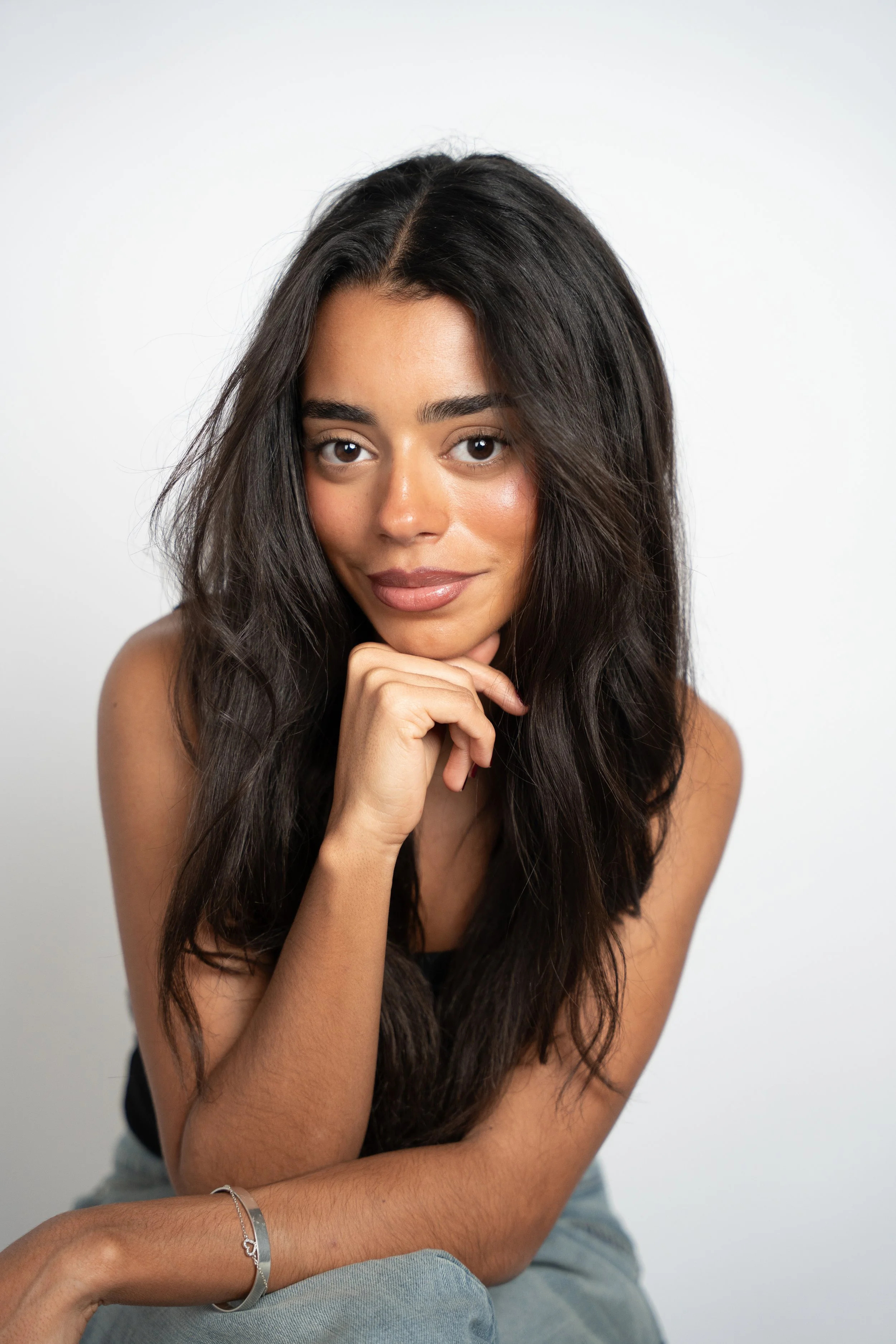 Portræt af en ung kvinde med langt, mørkt hår og et smil, der hviler sit hage i sin hånd, mod en hvid baggrund.