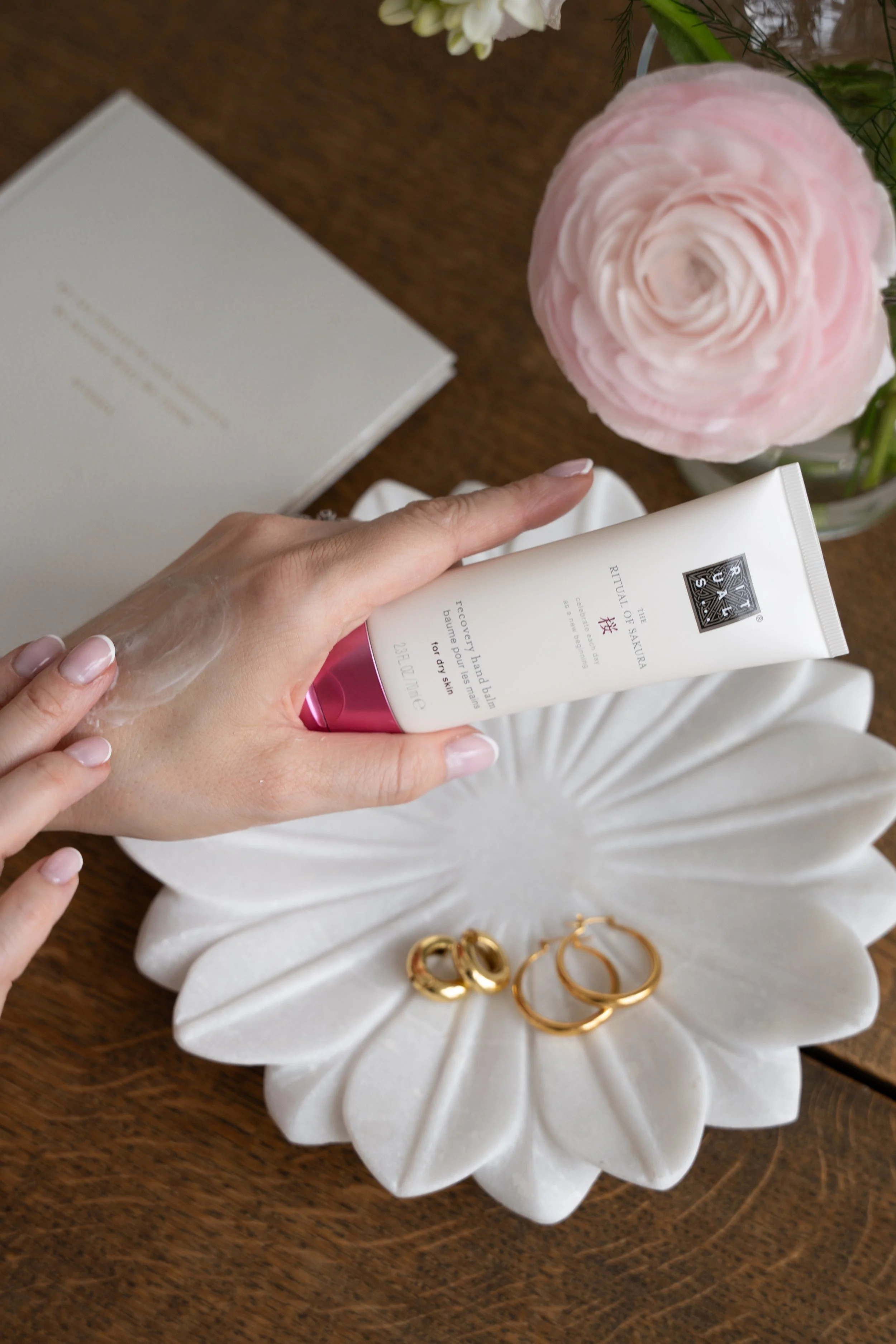 Person påfører ansigtspleje fra Rituals til huden, omgivet af guldøresmykker, en sorteret bog, en rosa ranunkelblomst og en gæstebog på et træbord.