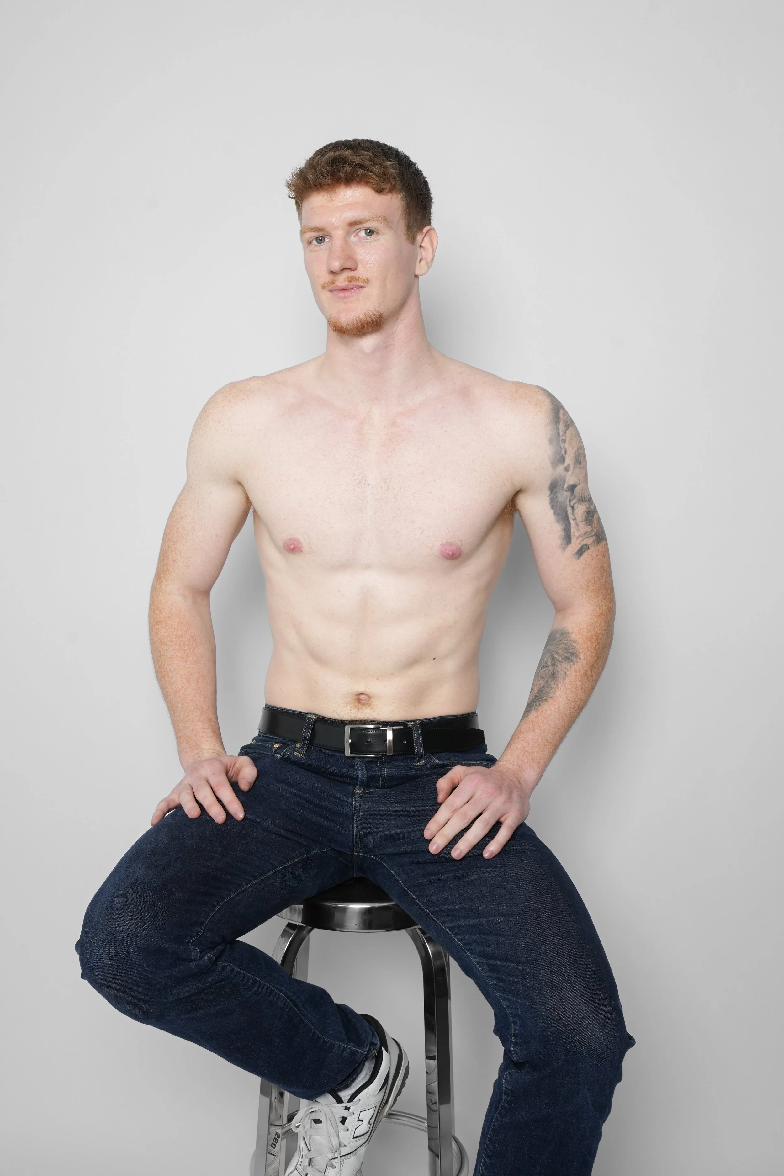 En shirtløs mand med auburn hår sidder på en sort barstol foran en hvid baggrund.