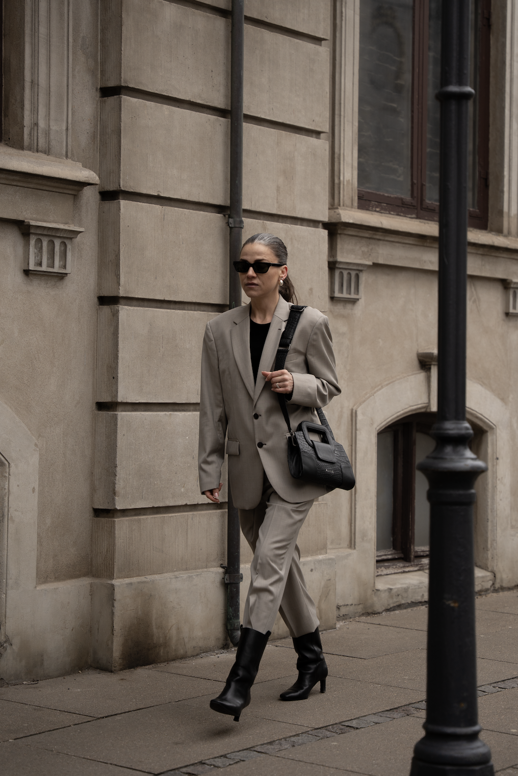 Kvinde i beige jakkesæt, sorte støvler og solbriller går på gaden ved en gammel bygning.
